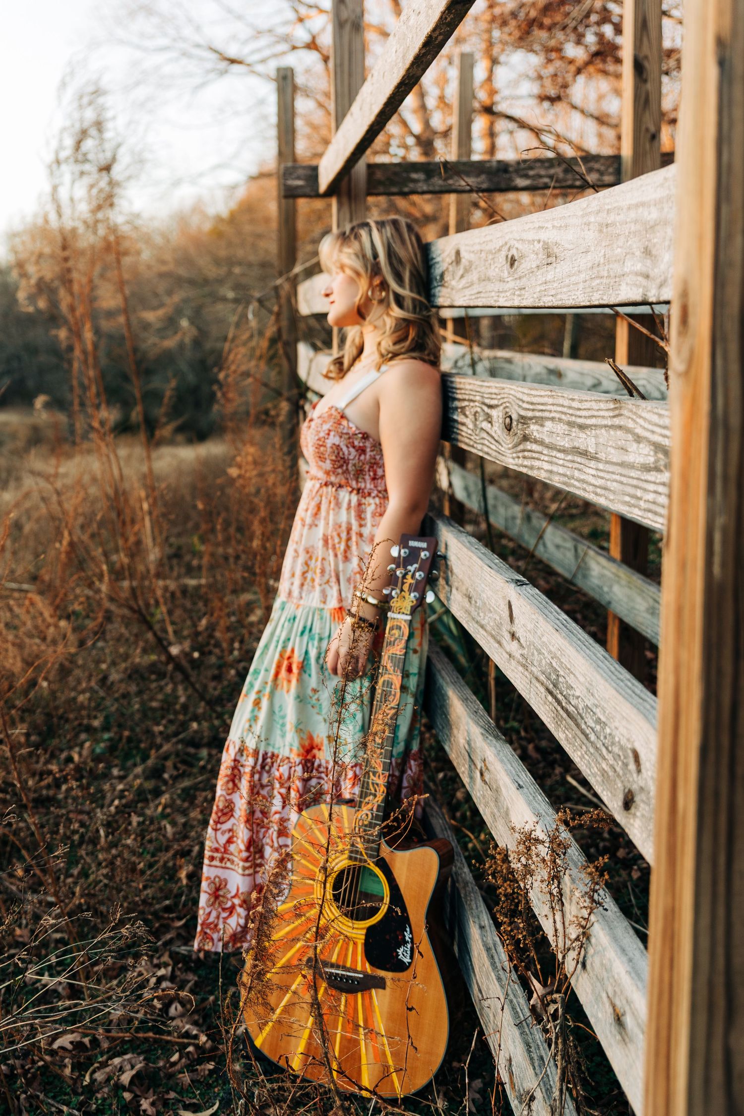 A person in a patterned maxi dress leans against a rustic wooden fence with an acoustic guitar at sunset.