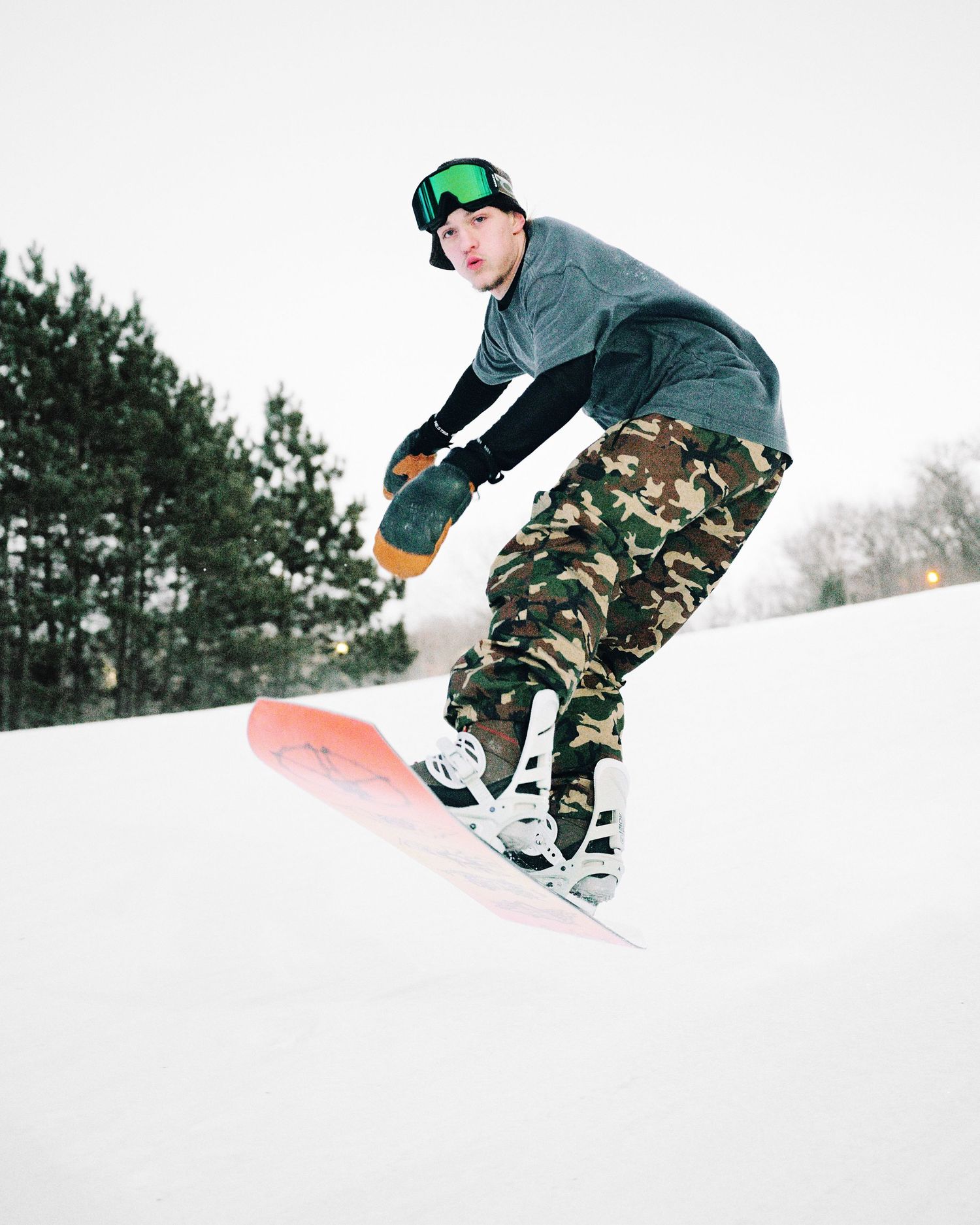 Snowboarder in camouflage pants and green goggles performing trick on orange board in snowy terrain.