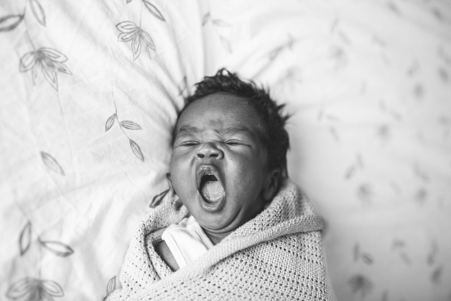 A sleepy newborn baby yawns while swaddled in a white knit blanket against floral patterned bedding.