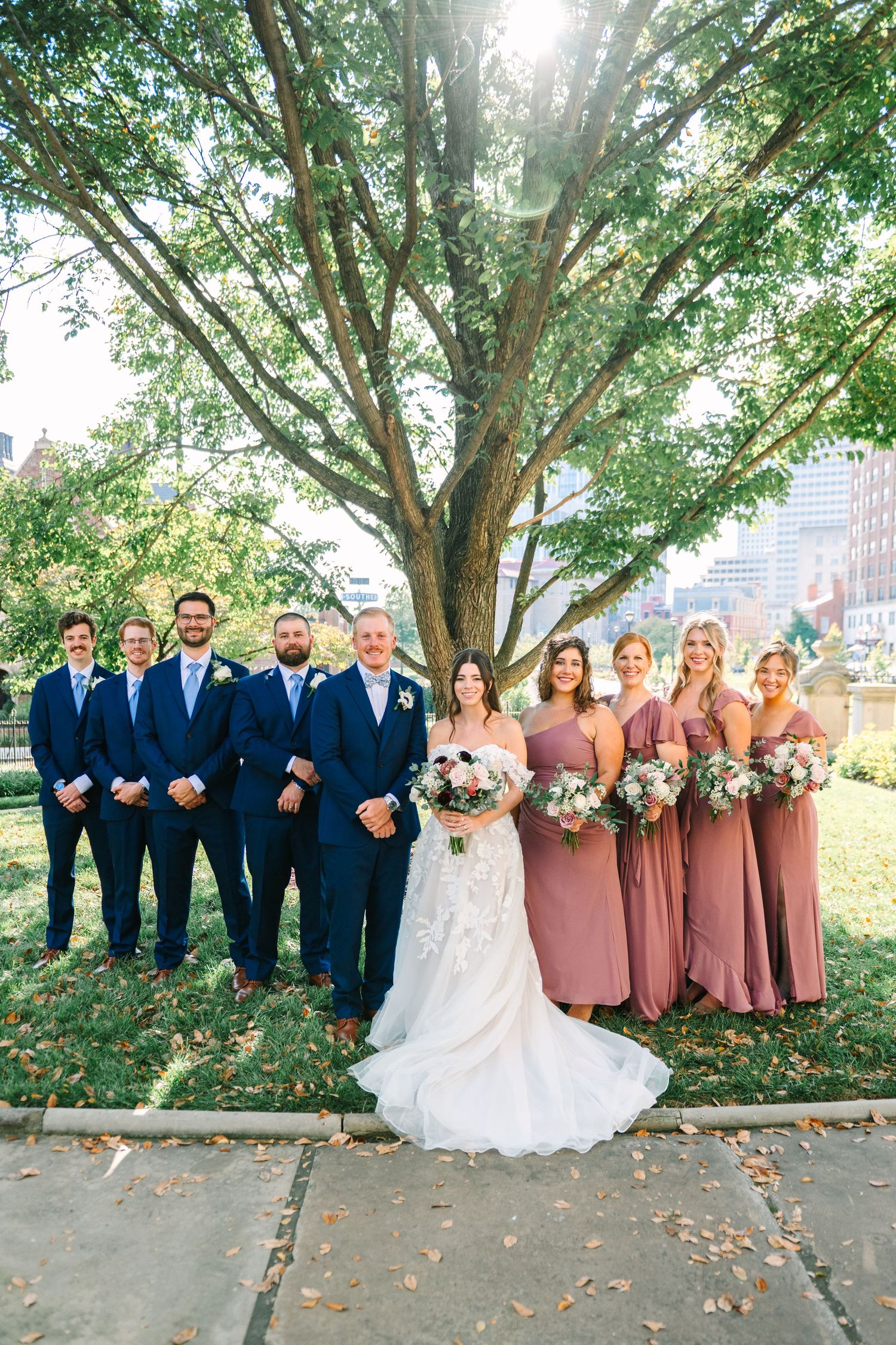 Wedding party in navy suits and dusty rose dresses poses together under trees on a fall day.