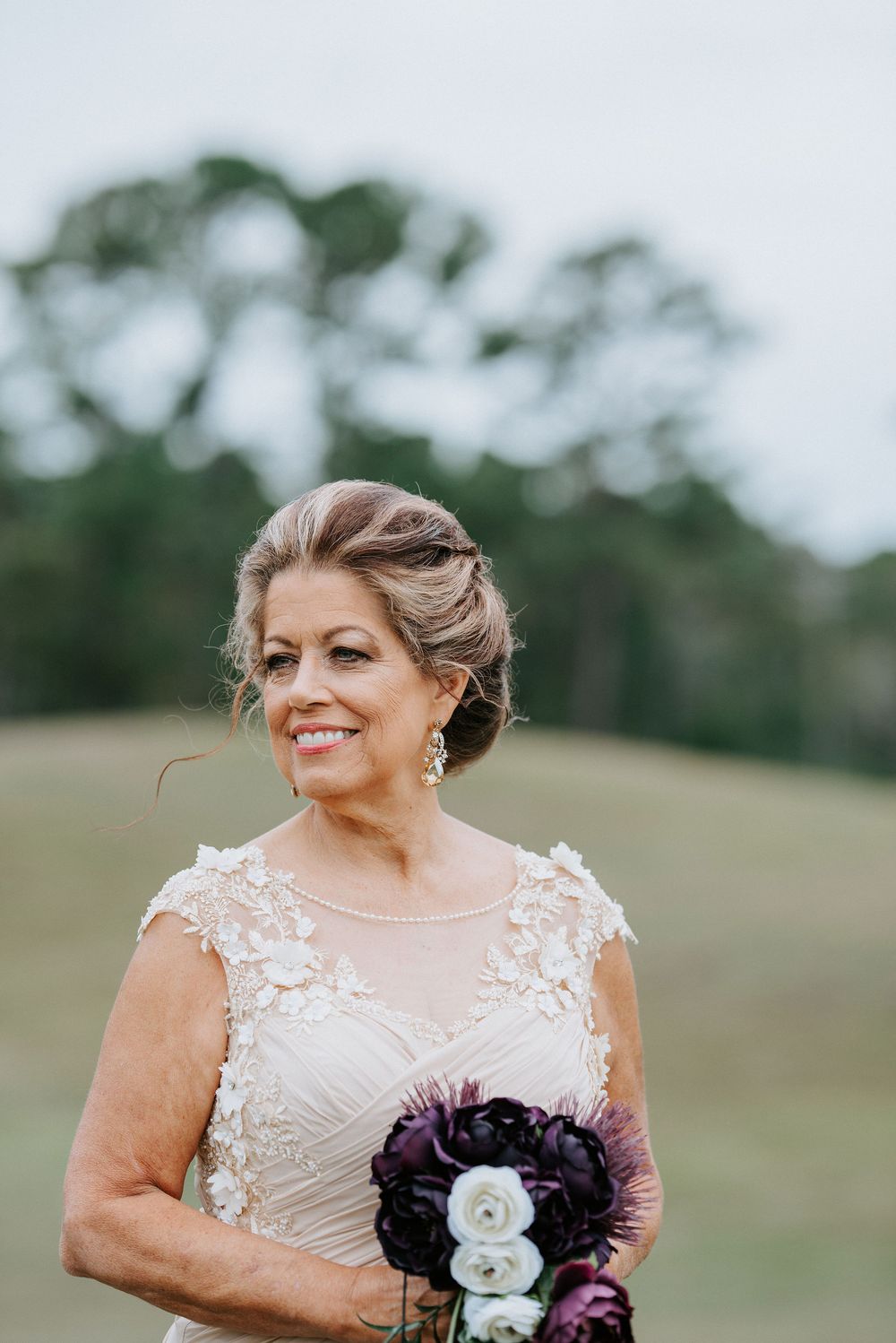 A bride in a white lace wedding dress holds a purple and white bouquet while standing in a grassy outdoor field.