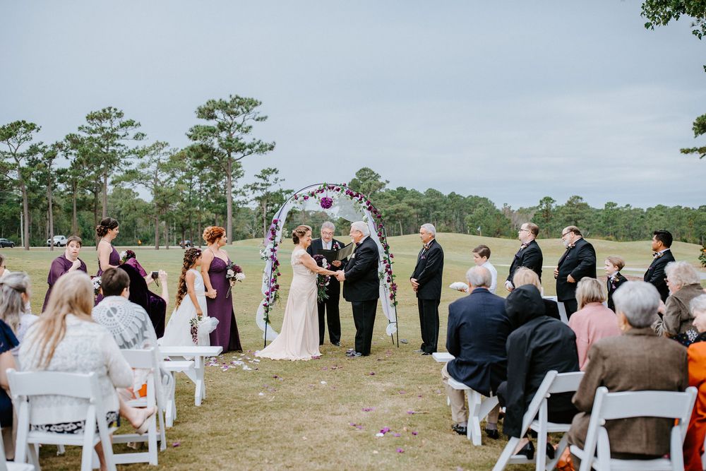 An outdoor wedding ceremony takes place in a scenic field with pine trees and white chairs set up for guests.