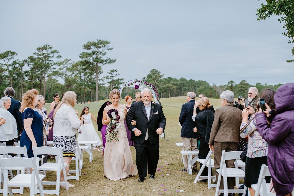 Wedding guests gather on white chairs during an outdoor ceremony with pine trees in the background.