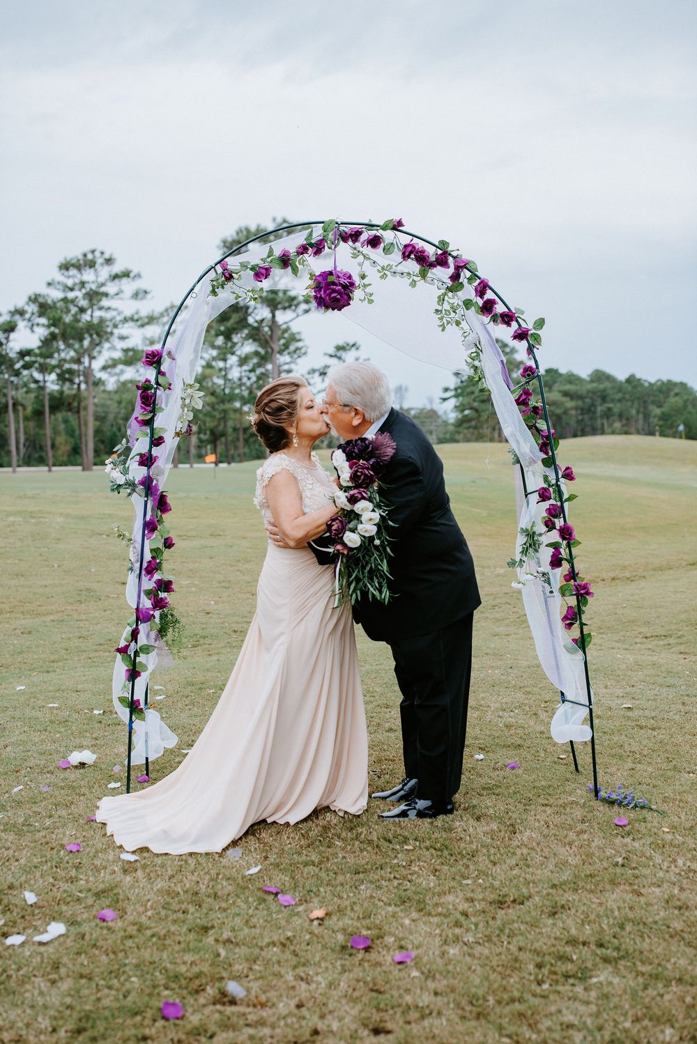 A romantic wedding ceremony under a floral arch decorated with purple and white flowers on a grassy hillside.