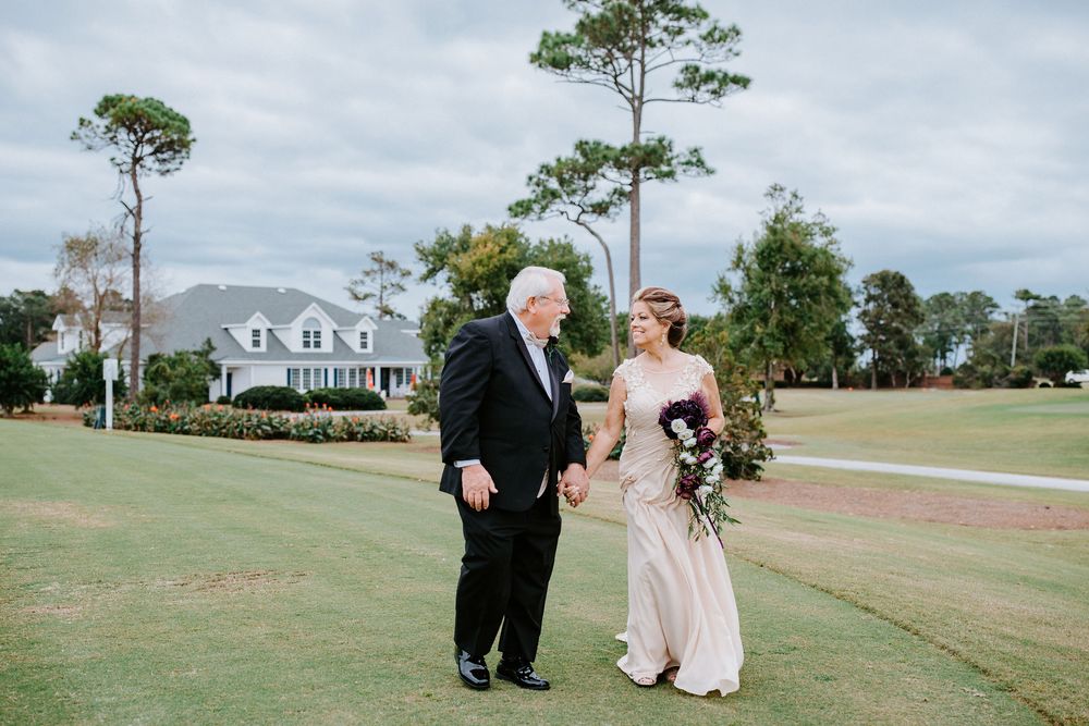 A couple shares a tender moment on a lush golf course with tall pine trees and a white house in the background.