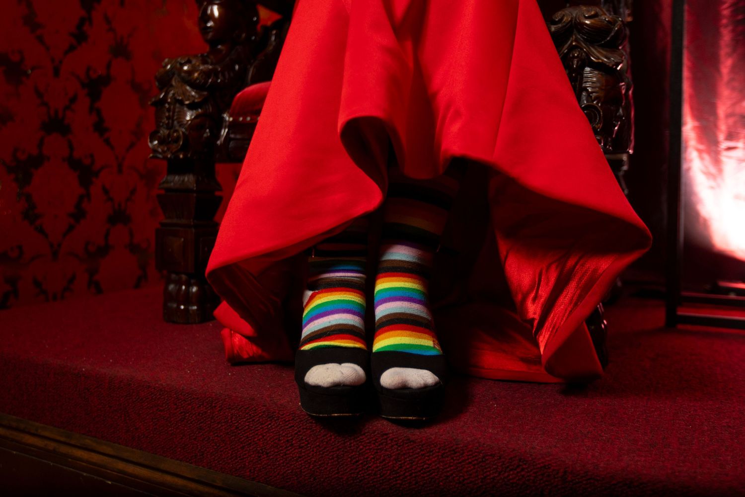A bride wears colorful striped socks in black high heels that peek out from under a flowing red dress in a dimly lit room with ornate decor.