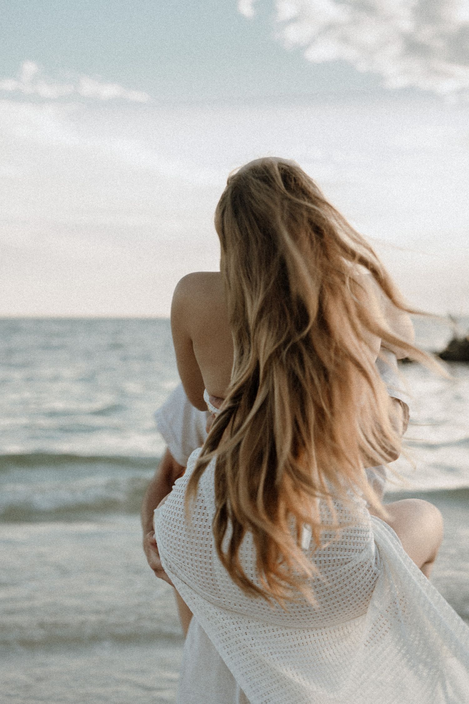 Woman with windswept blonde hair in white skirt and bikini looks out at ocean waves during golden hour at beach engagement session