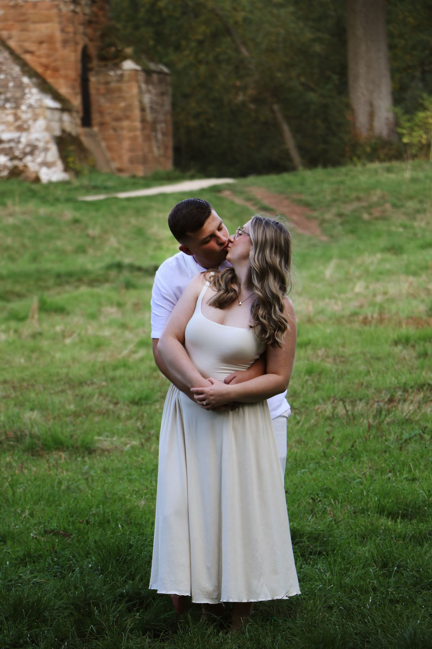 A couple in light-colored outfits share a romantic embrace on a grassy lawn near an old brick wall.