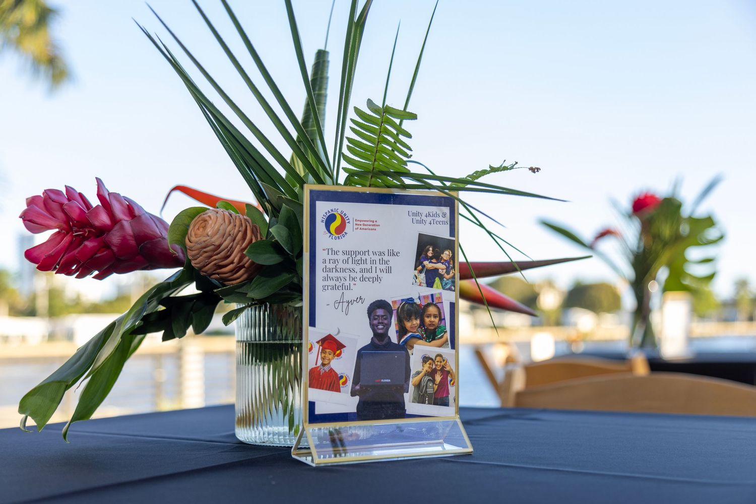 Tropical floral centerpiece with pink ginger and palm leaves beside a framed photo display on an outdoor event table.