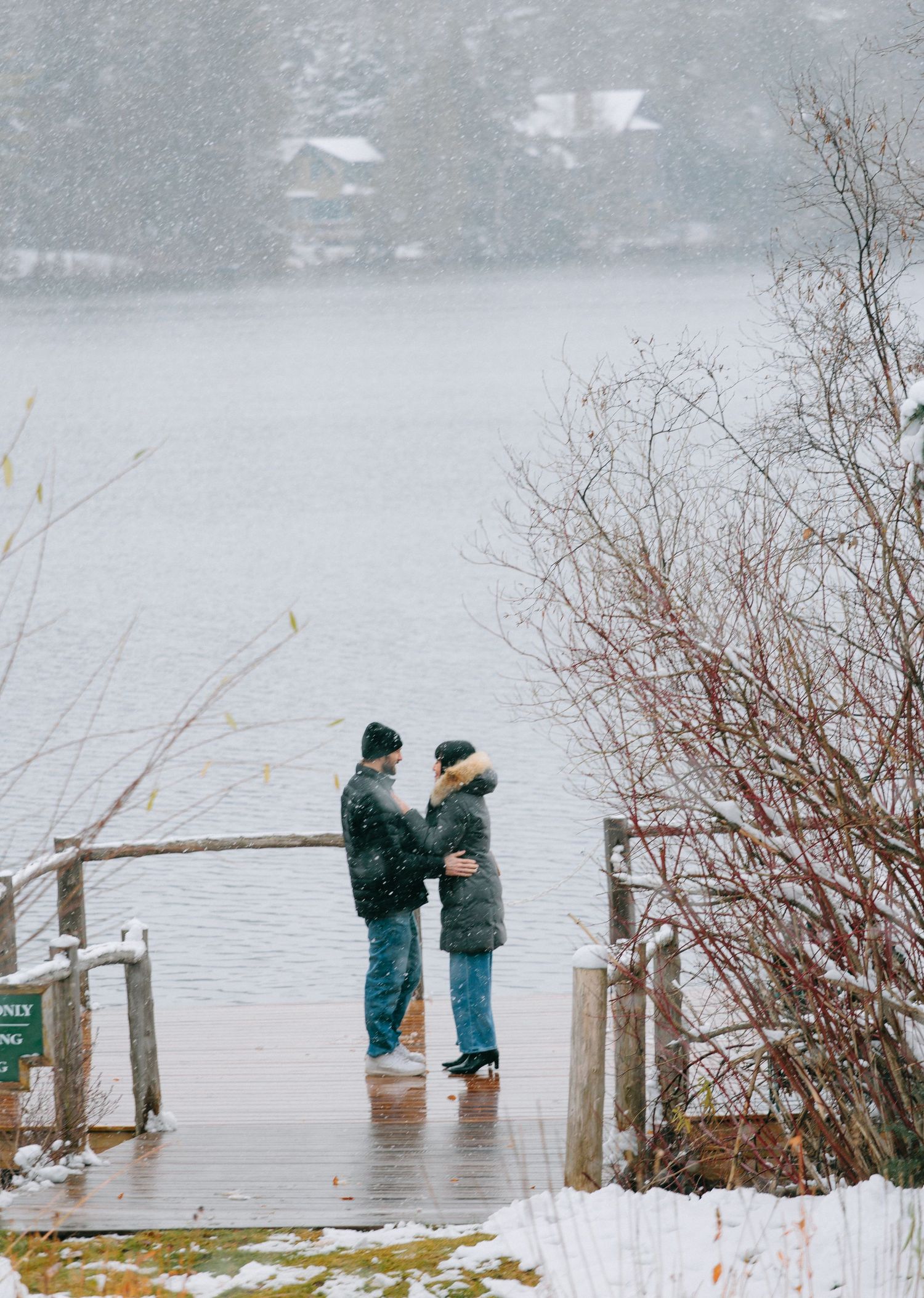 Two people stand on a snowy dock overlooking a misty lake on a winter day.