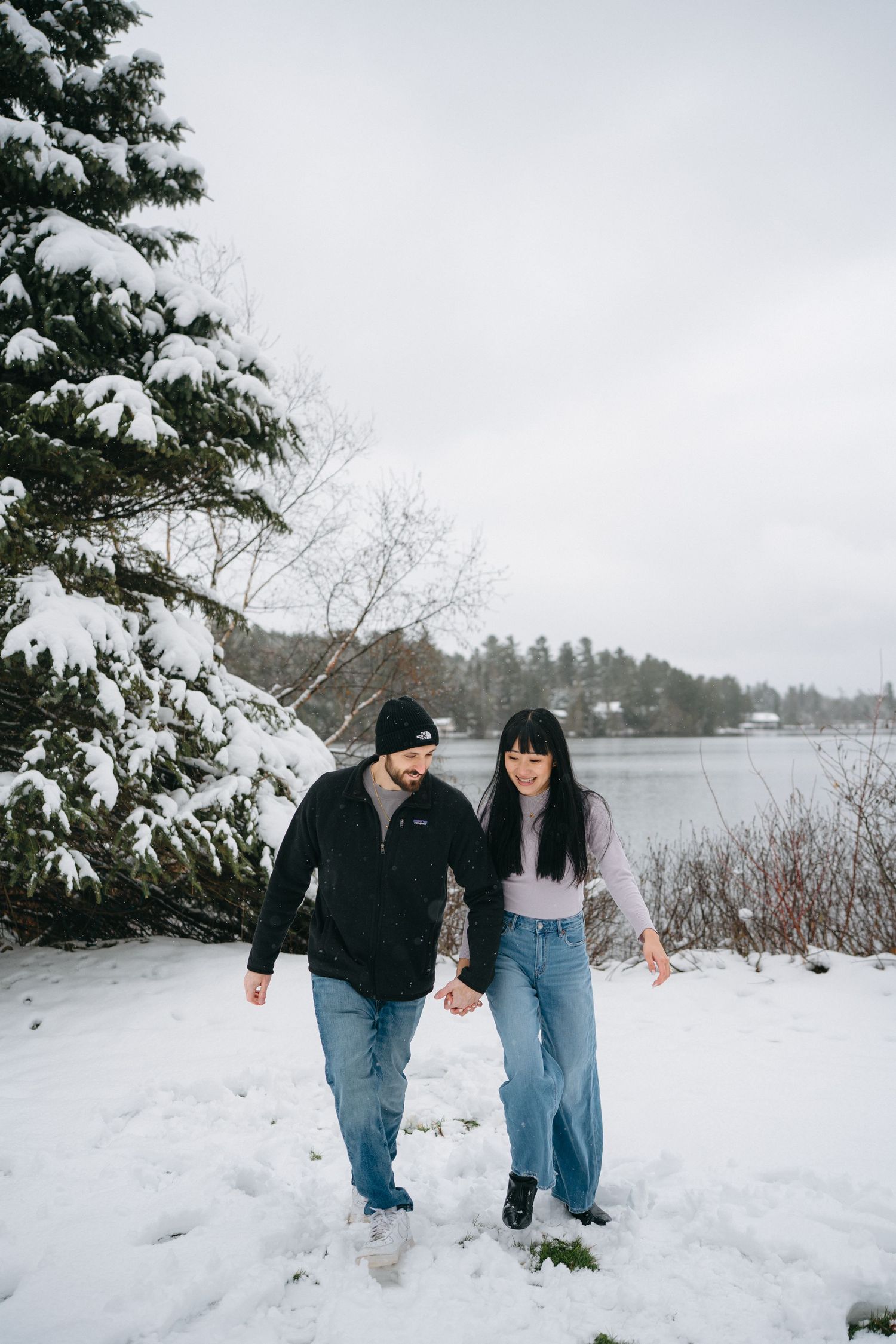 A couple walks hand in hand through a snow-covered landscape with evergreen trees and a lake in the background.
