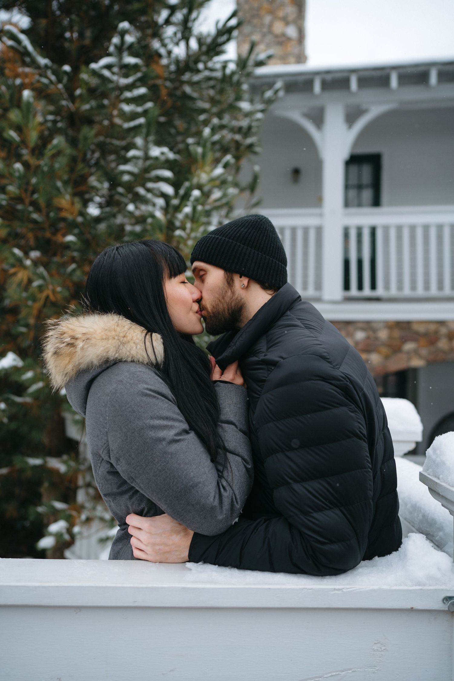 A romantic winter kiss shared on a white porch railing with snow-covered scenery in the background.