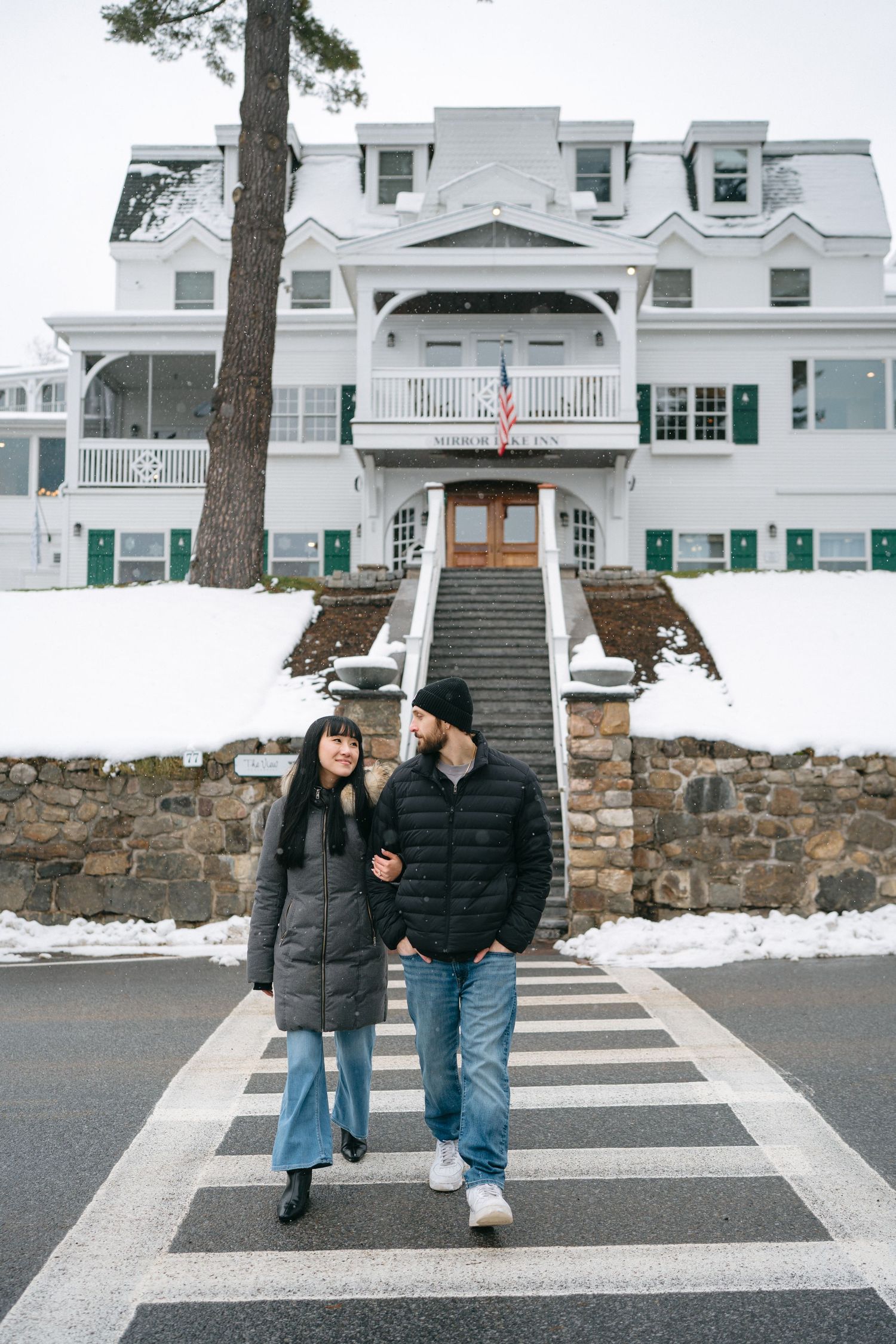 A couple in winter coats walking across a crosswalk in front of a grand white colonial building in the snow.