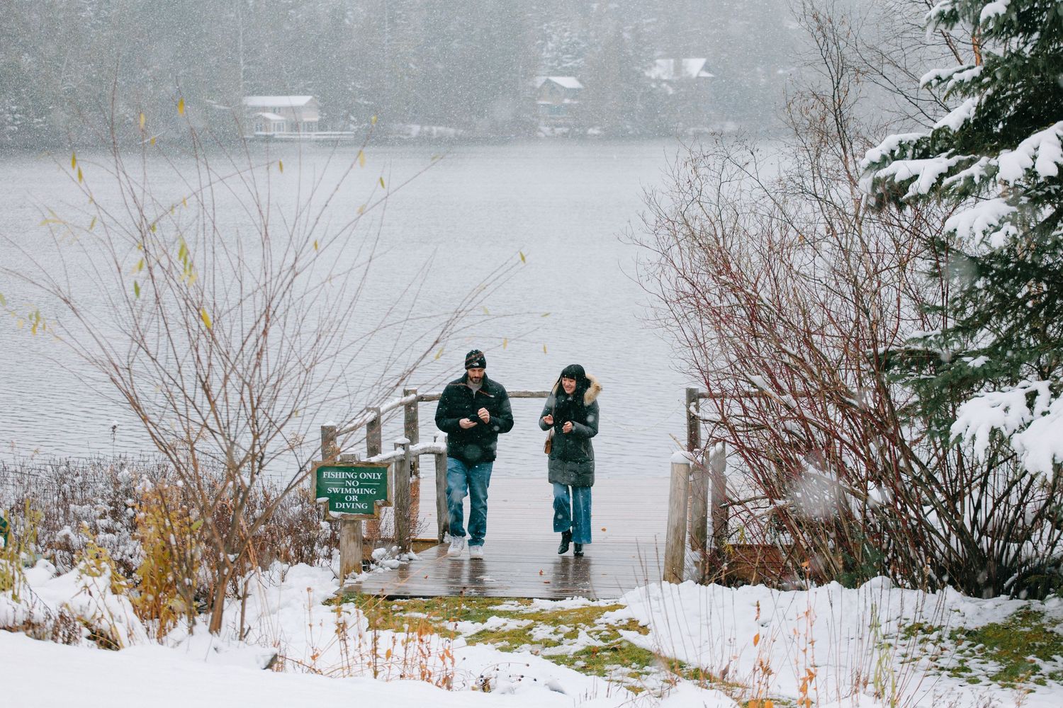 Two people stand on a snowy dock overlooking a misty lake in winter with bare trees and snow-covered ground.