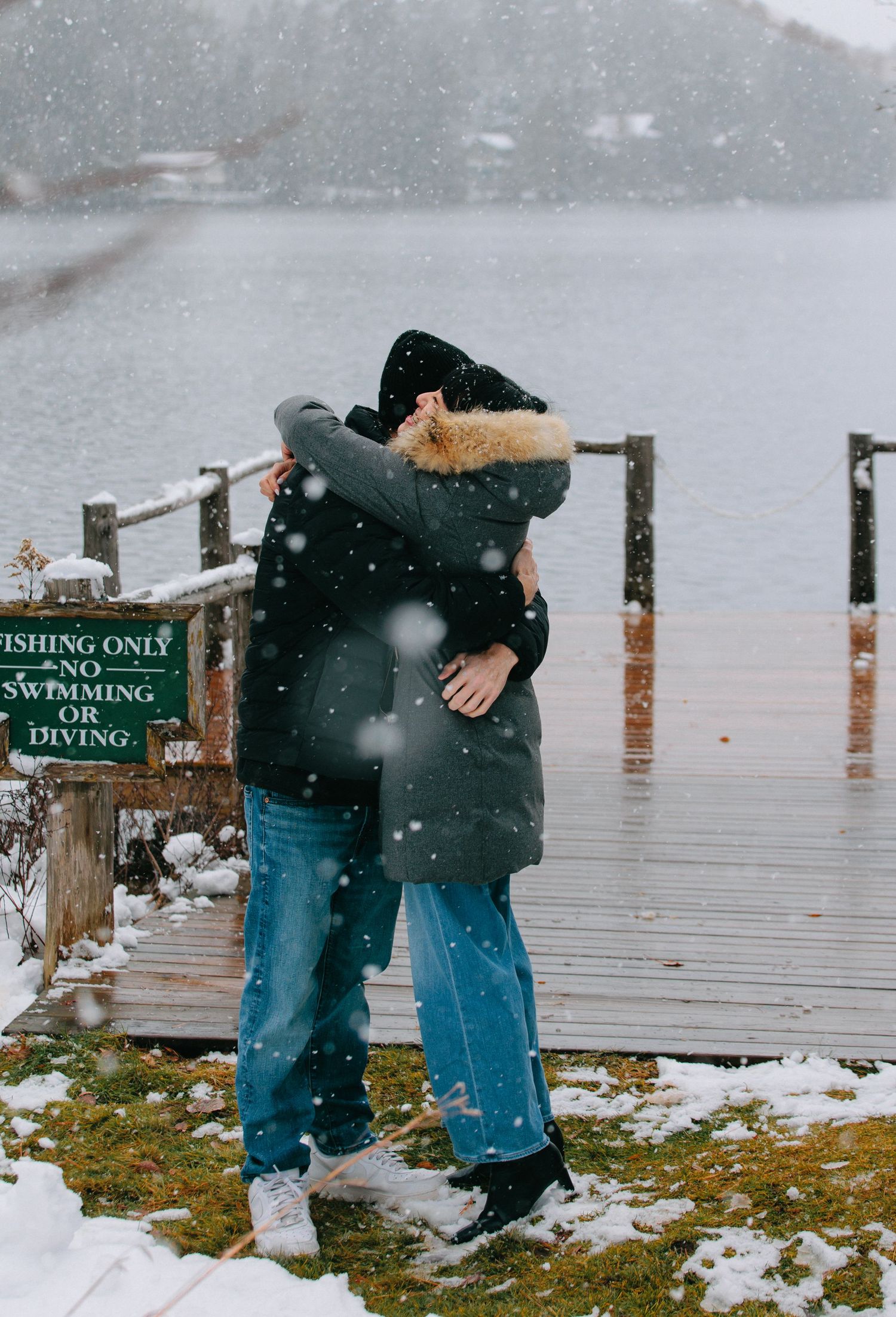A couple playfully poses on a wooden dock with snow-covered signs in winter.