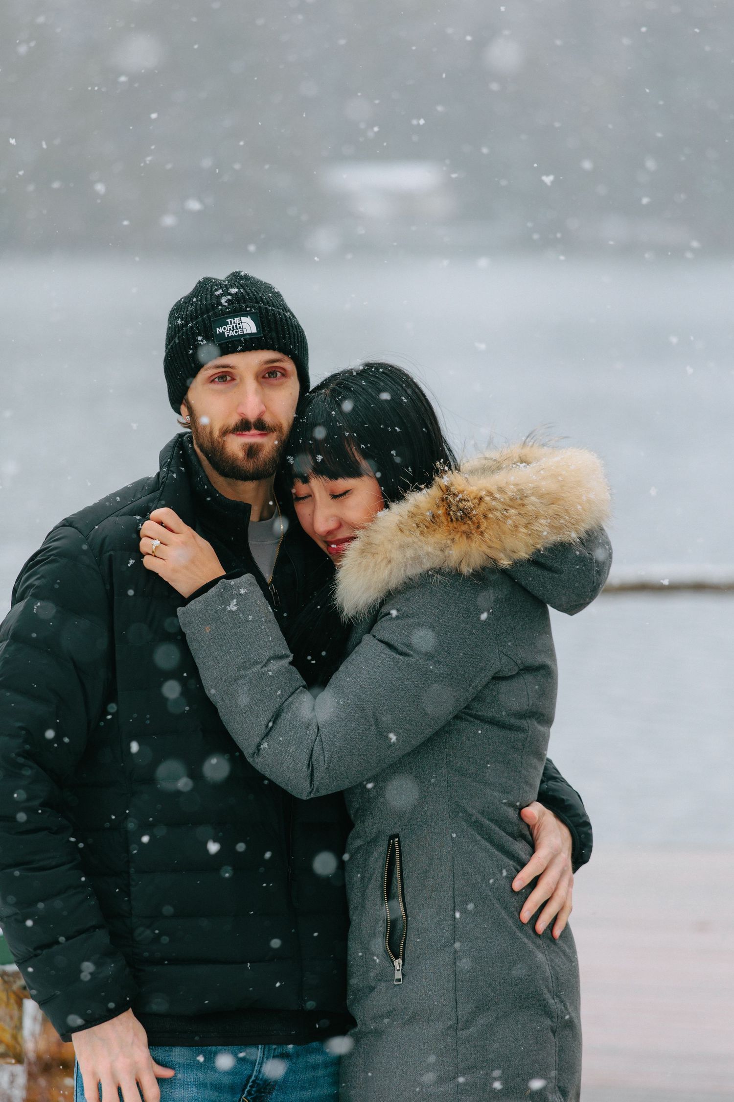 A couple embraces in winter snowfall wearing warm coats and beanies.