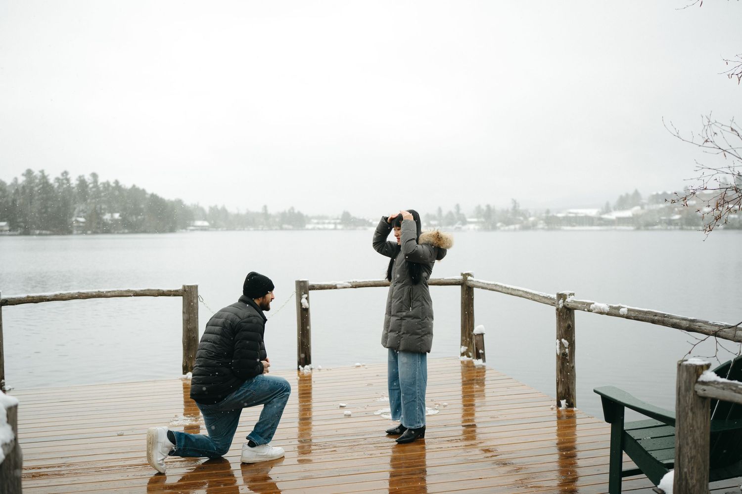Two people stand on a snowy winter dock overlooking a misty lake with trees in the background.