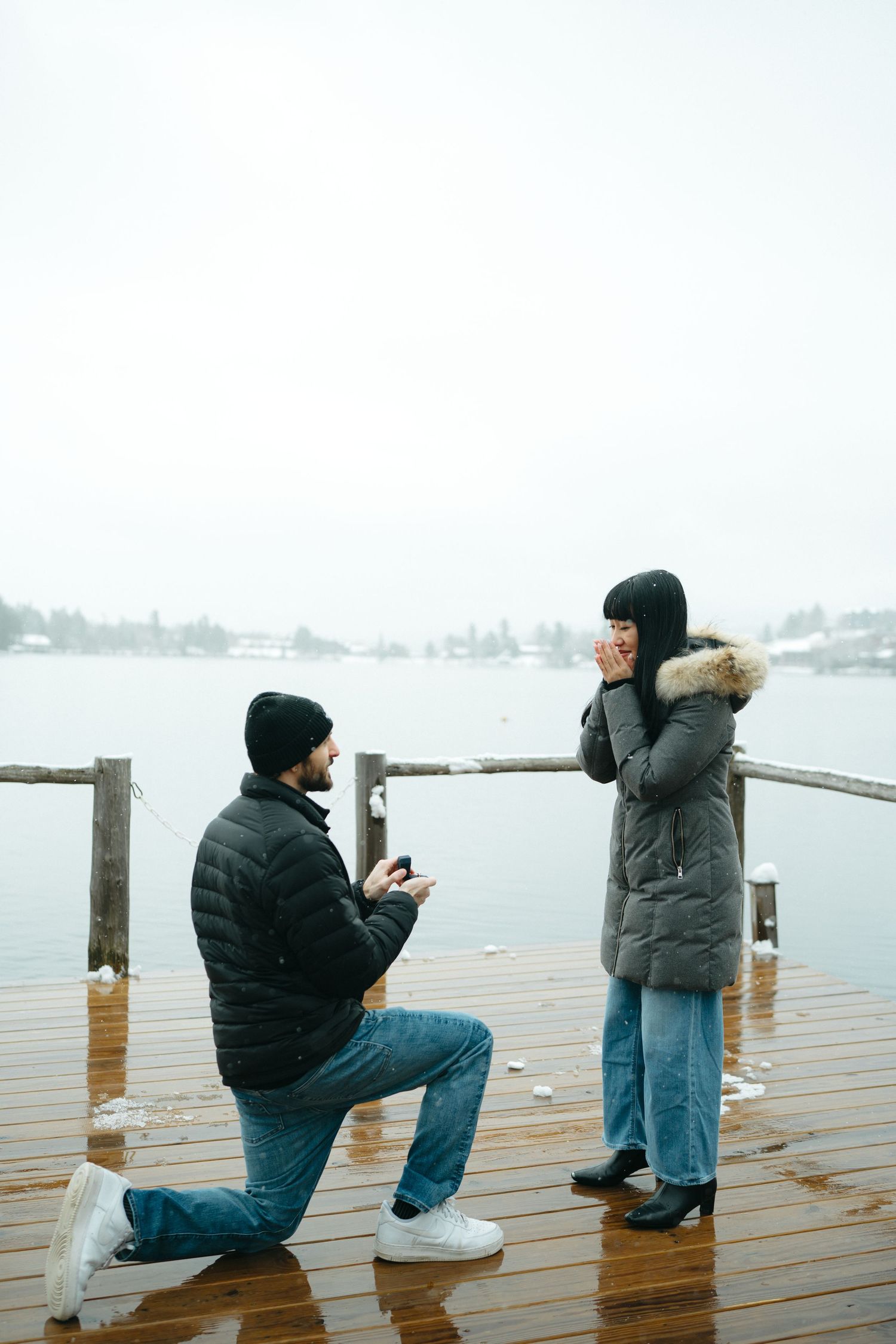 A romantic marriage proposal takes place on a wet wooden dock overlooking a misty lake on a winter day.