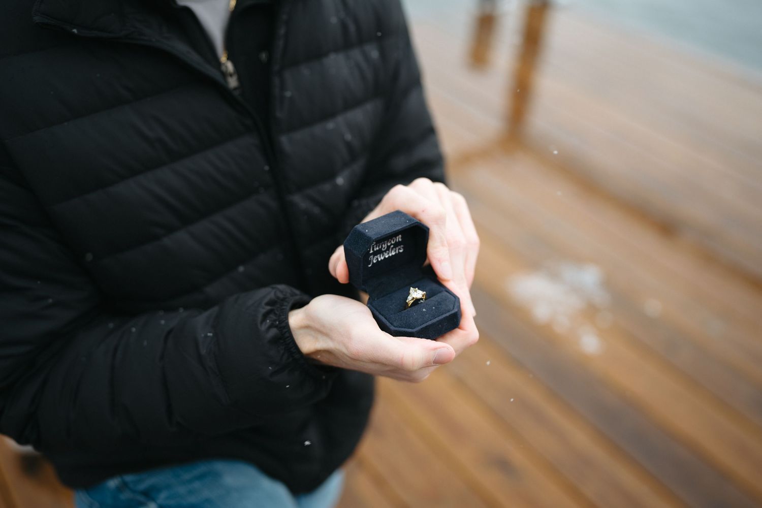 A close-up shot of hands holding a small navy blue ring box on a wooden deck.