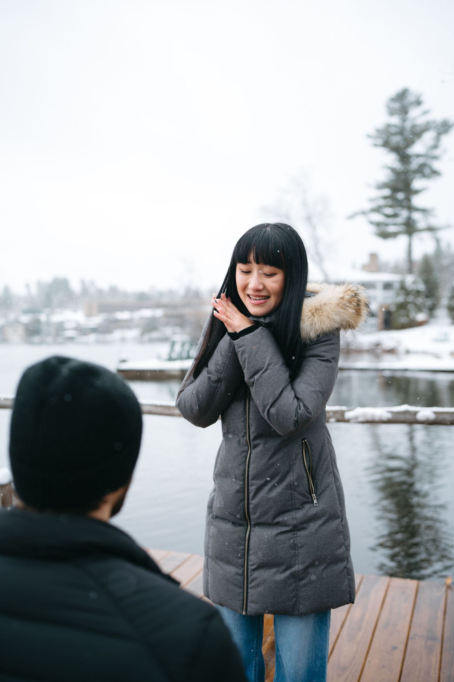 A joyful winter proposal scene on a snowy dock with pine trees in the background.