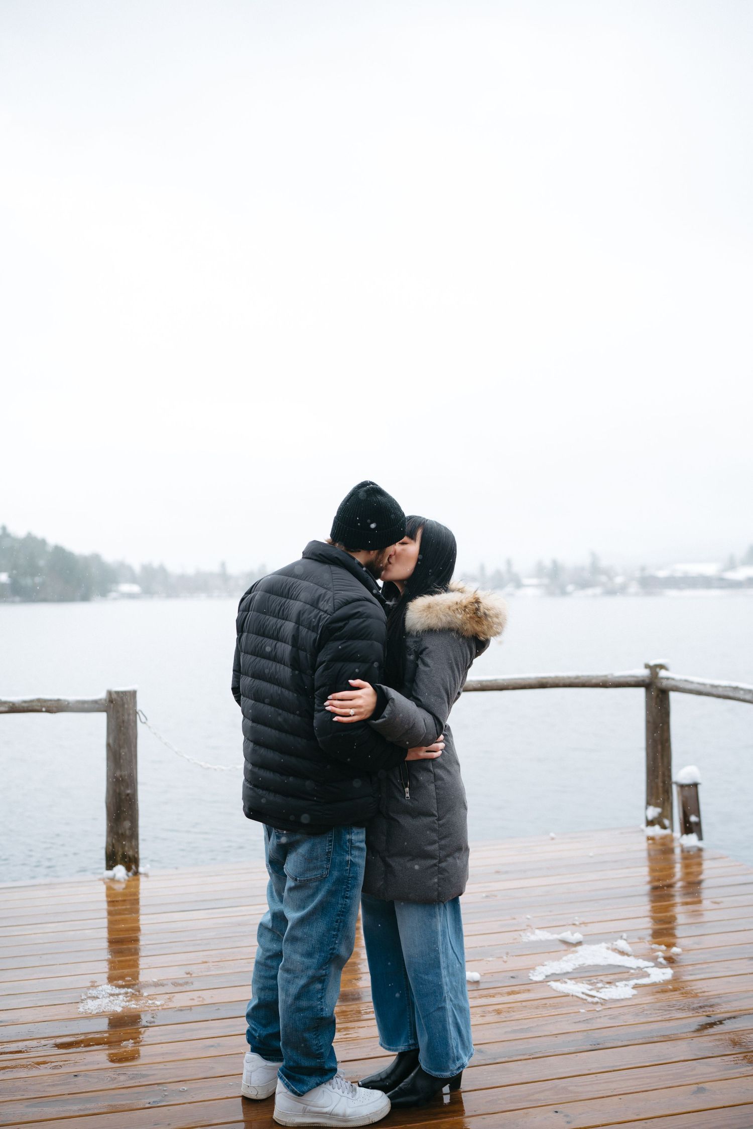 A couple embraces on a rainy wooden dock with a foggy lake view in the background.