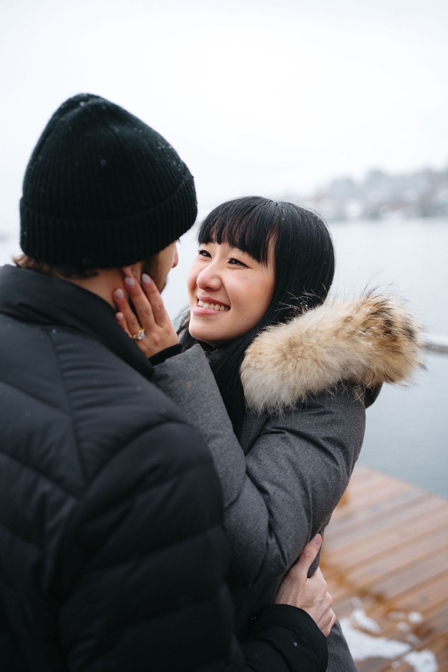 A couple sharing an intimate winter moment outdoors by a lake, wearing warm coats and smiling at each other.