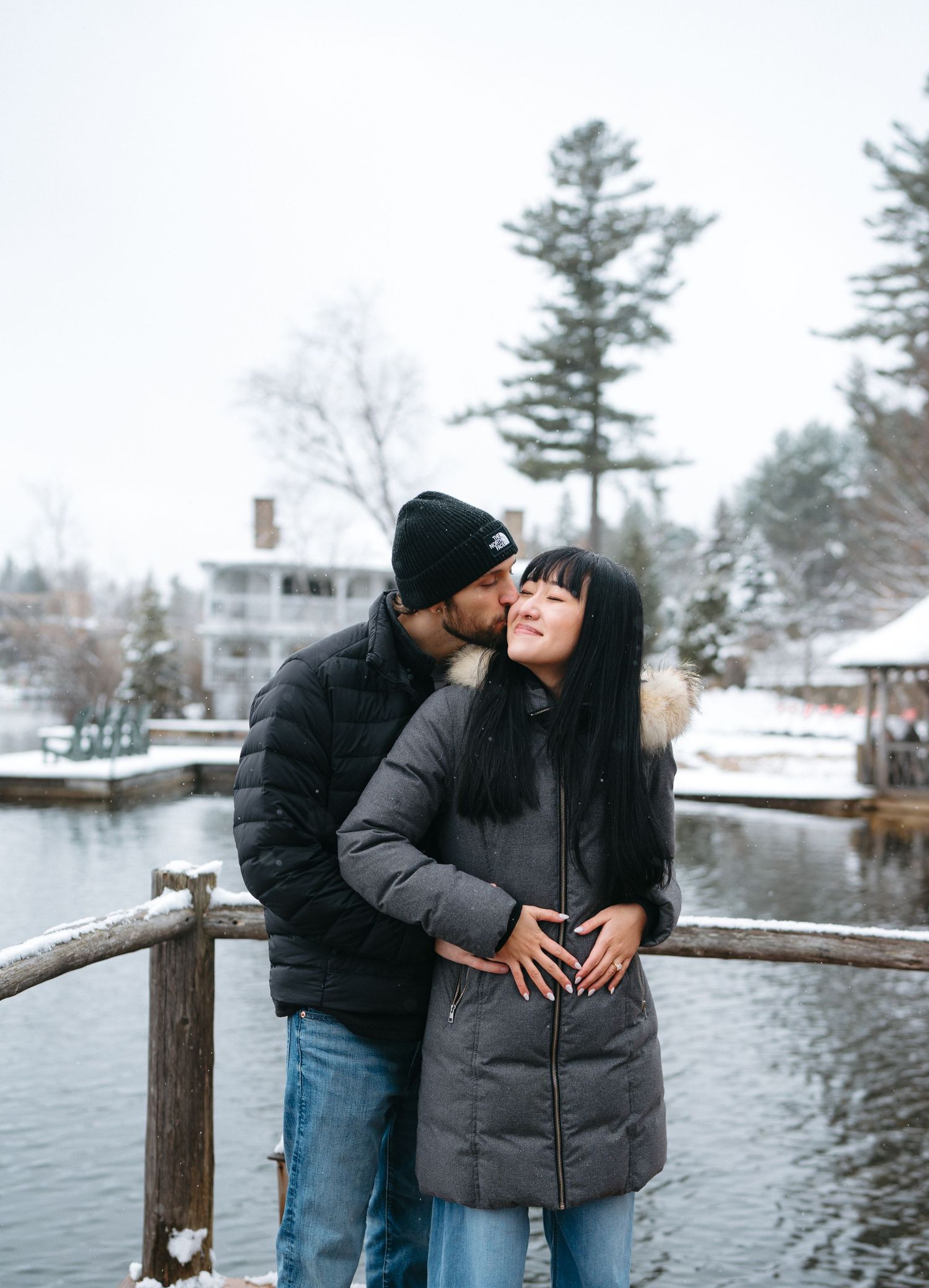 A couple shares an intimate moment on a wooden railing by a snowy lake with evergreen trees in the background.