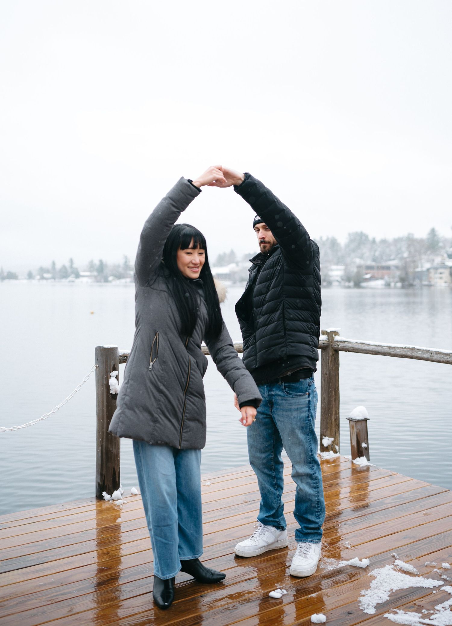 Two people dancing and laughing together on a wooden dock overlooking a winter lake.