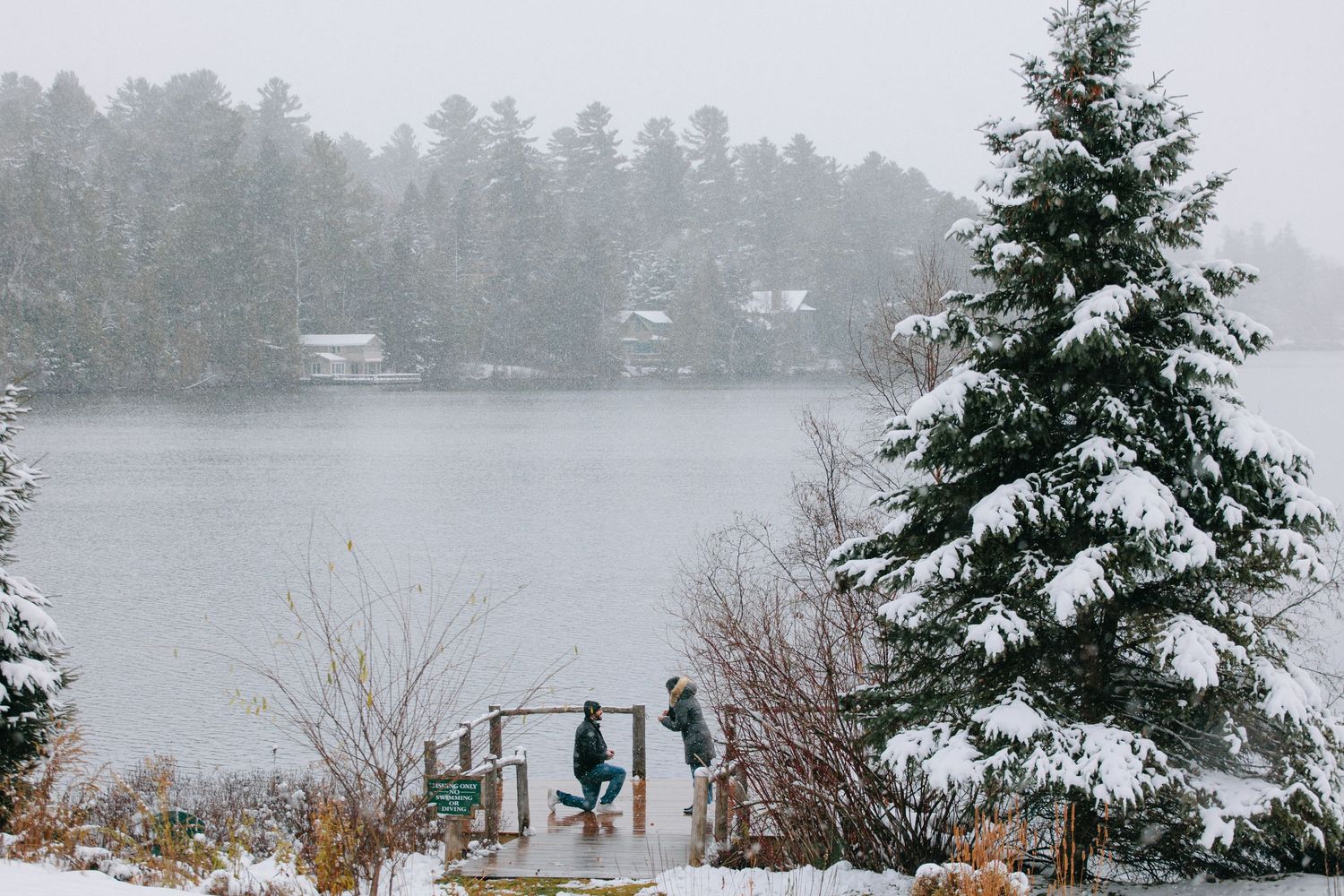 Two people stand on a snowy dock overlooking a misty lake with evergreen trees and houses along the shoreline.