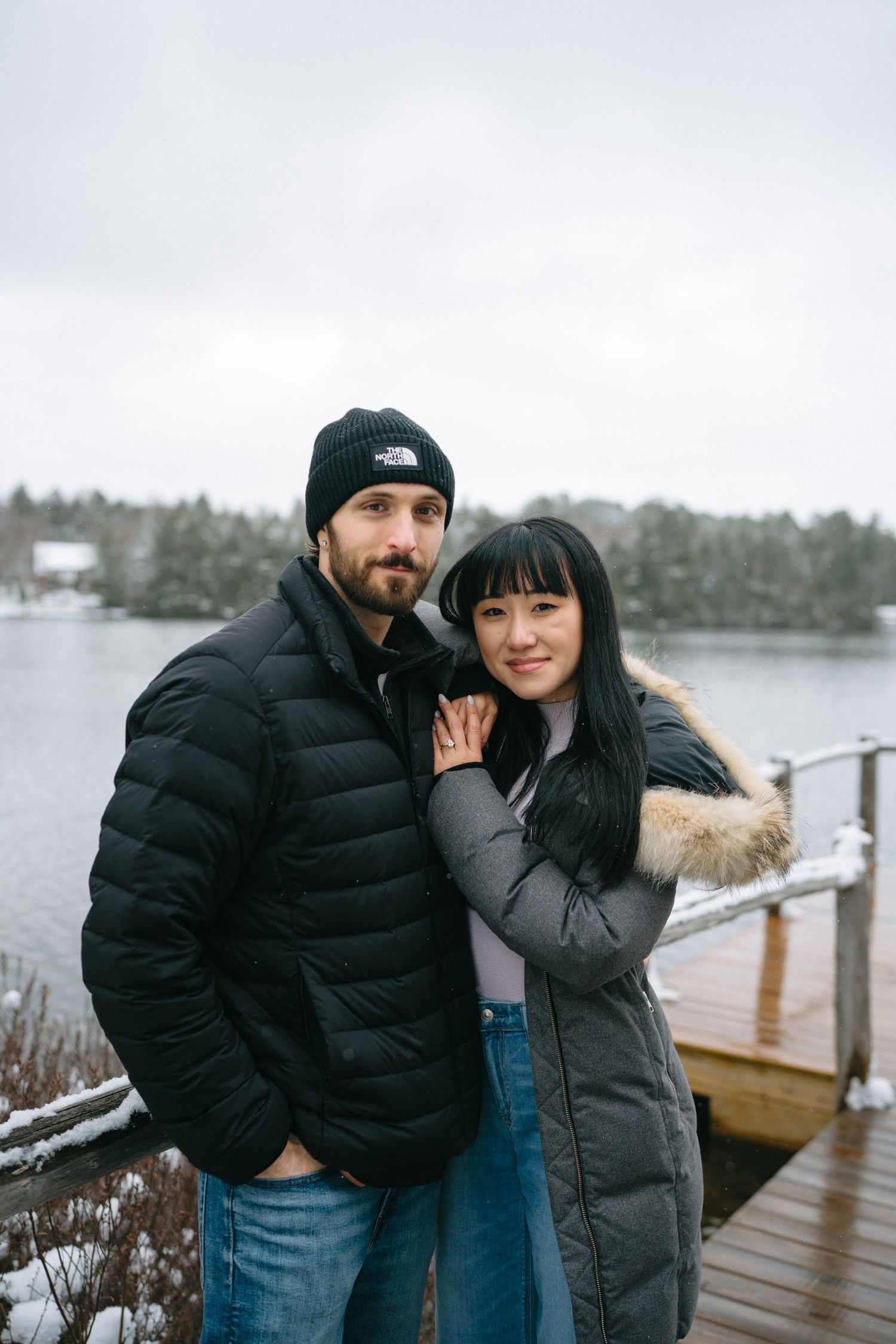 A couple in winter coats share intimate moments on a snowy dock by a lake.