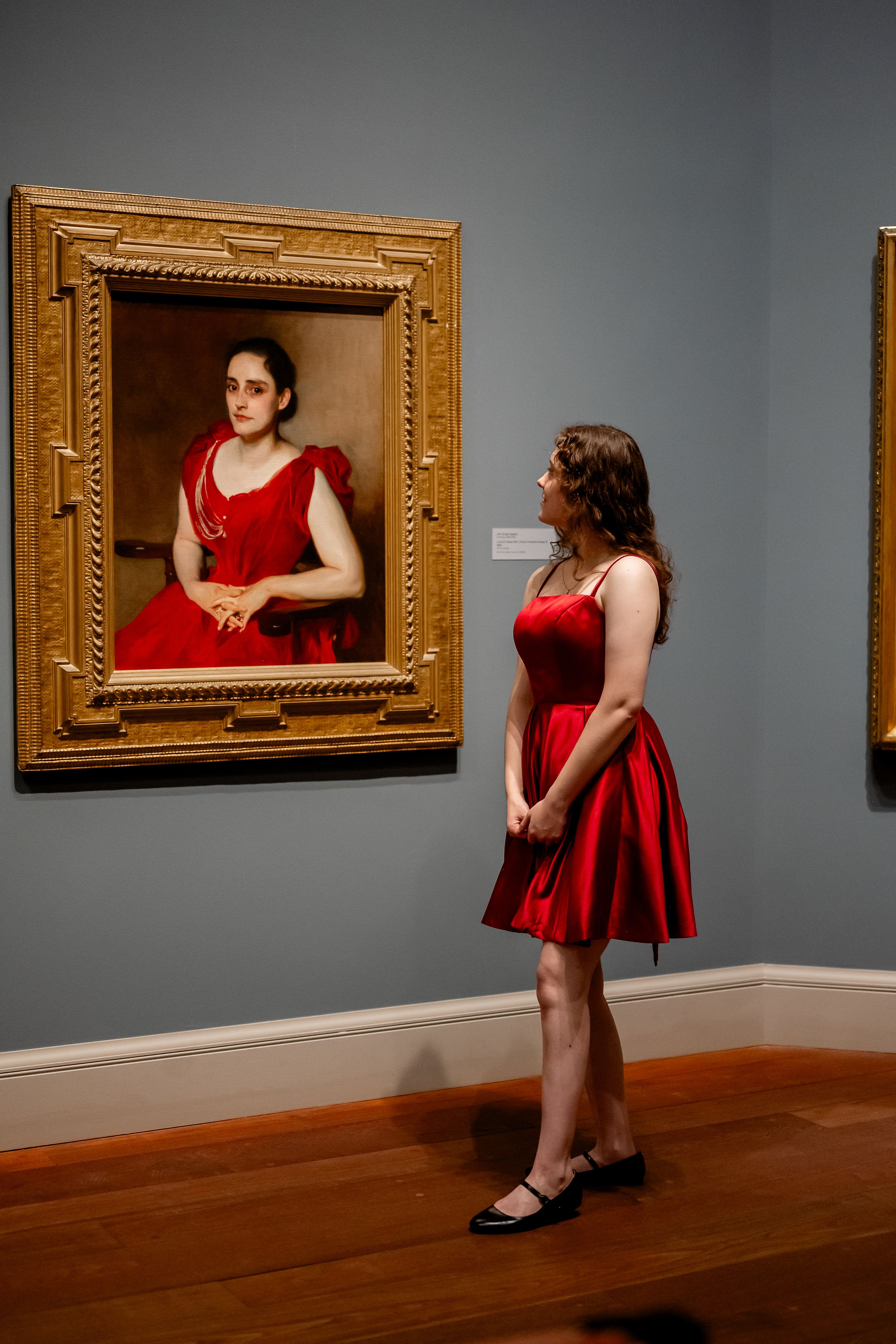 A person in a red dress stands in an art gallery viewing a portrait painting in an ornate gold frame.