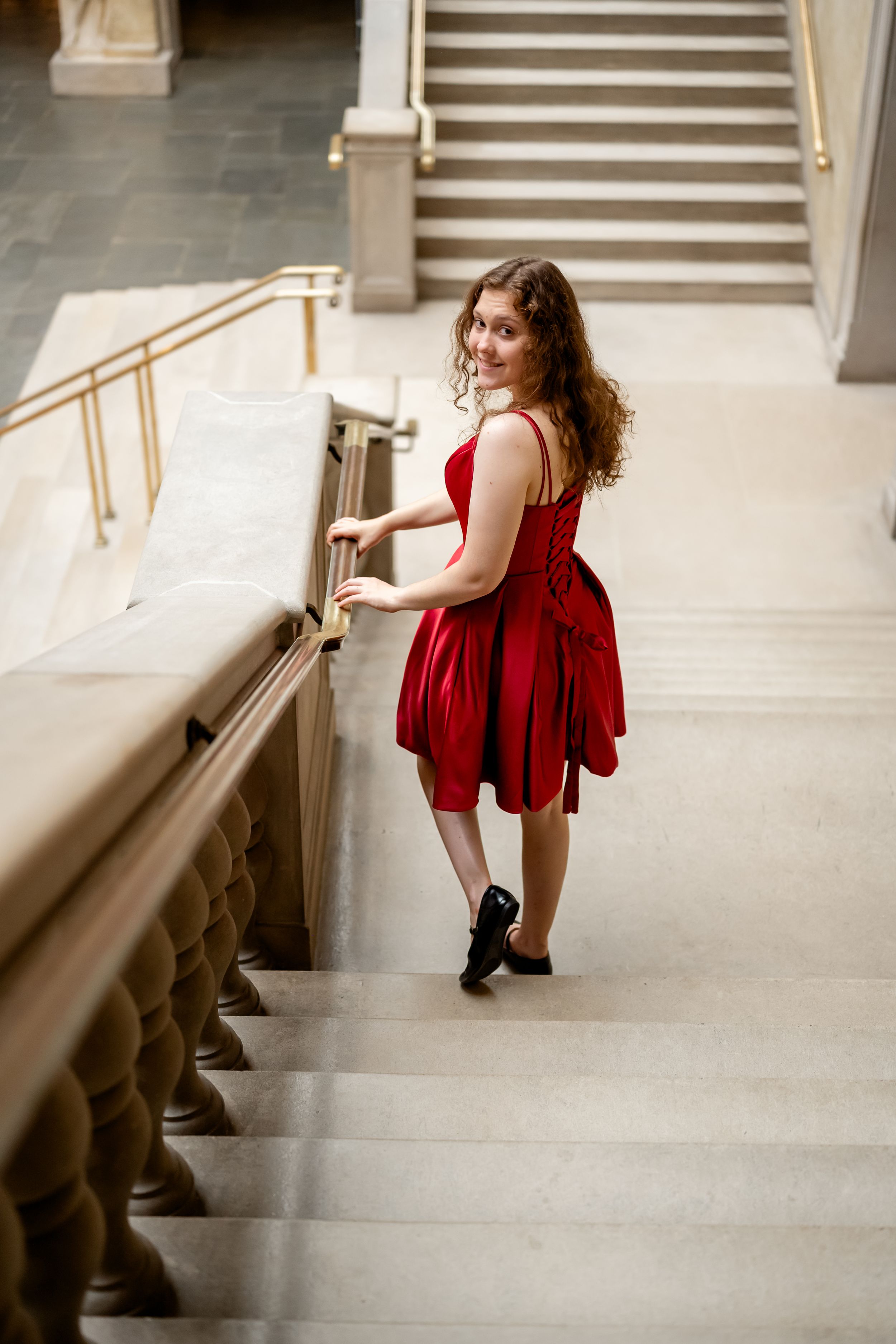 A figure in a flowing red dress poses gracefully on marble stairs in an elegant architectural setting.