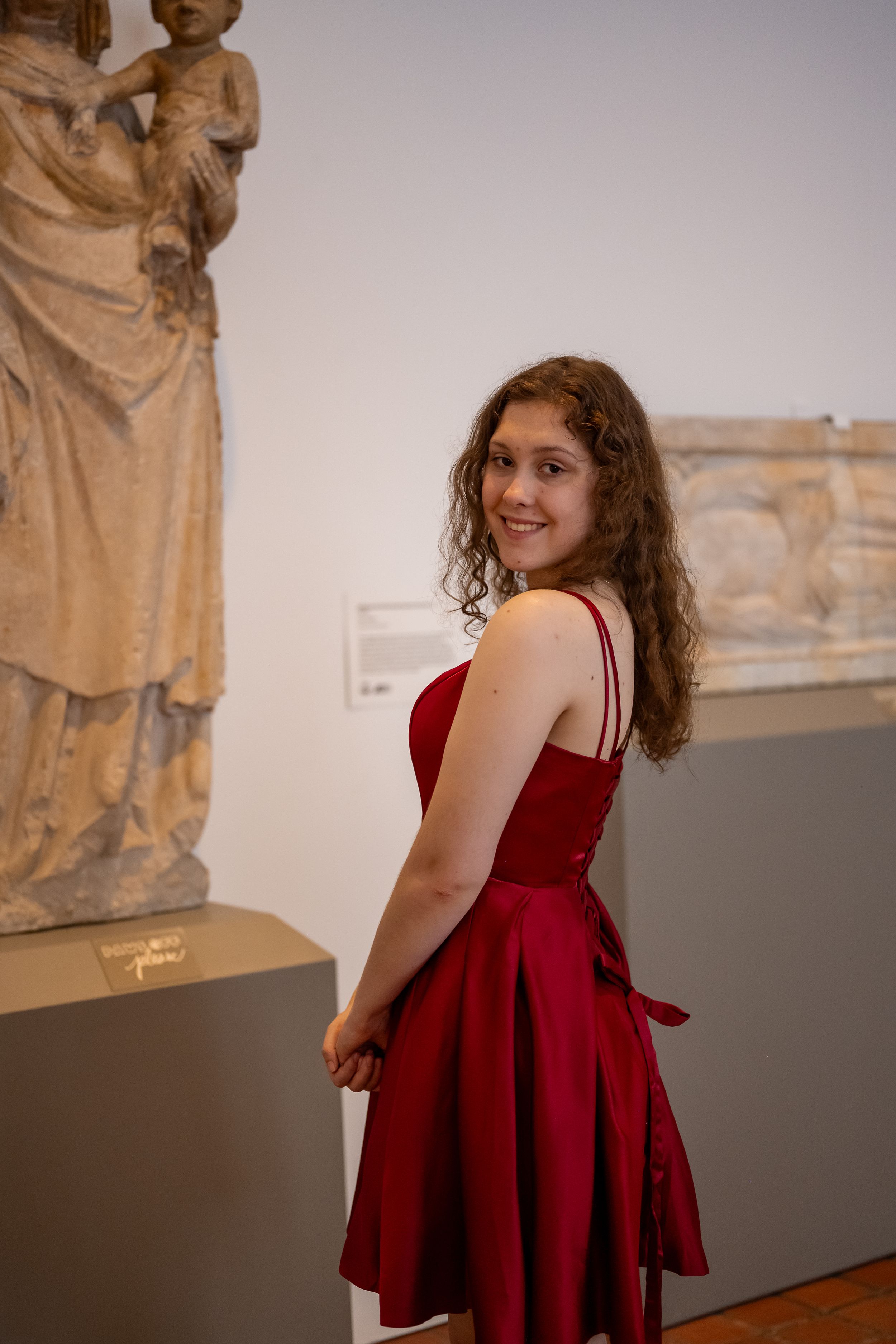 A person wearing a red dress stands among classical sculptures in a museum gallery space.
