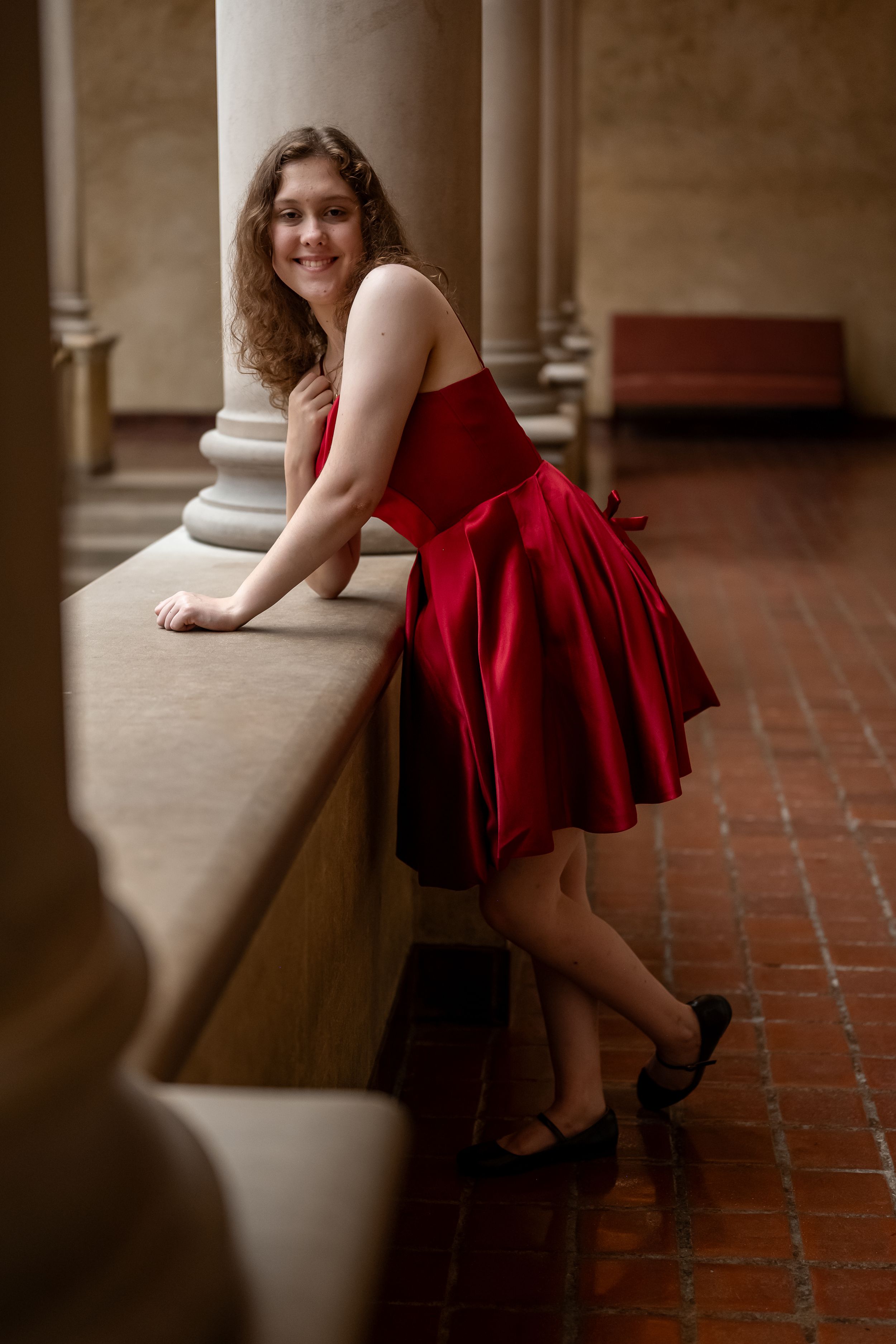 A woman in a flowing red dress and heels leans against a stone ledge in an elegant architectural setting.