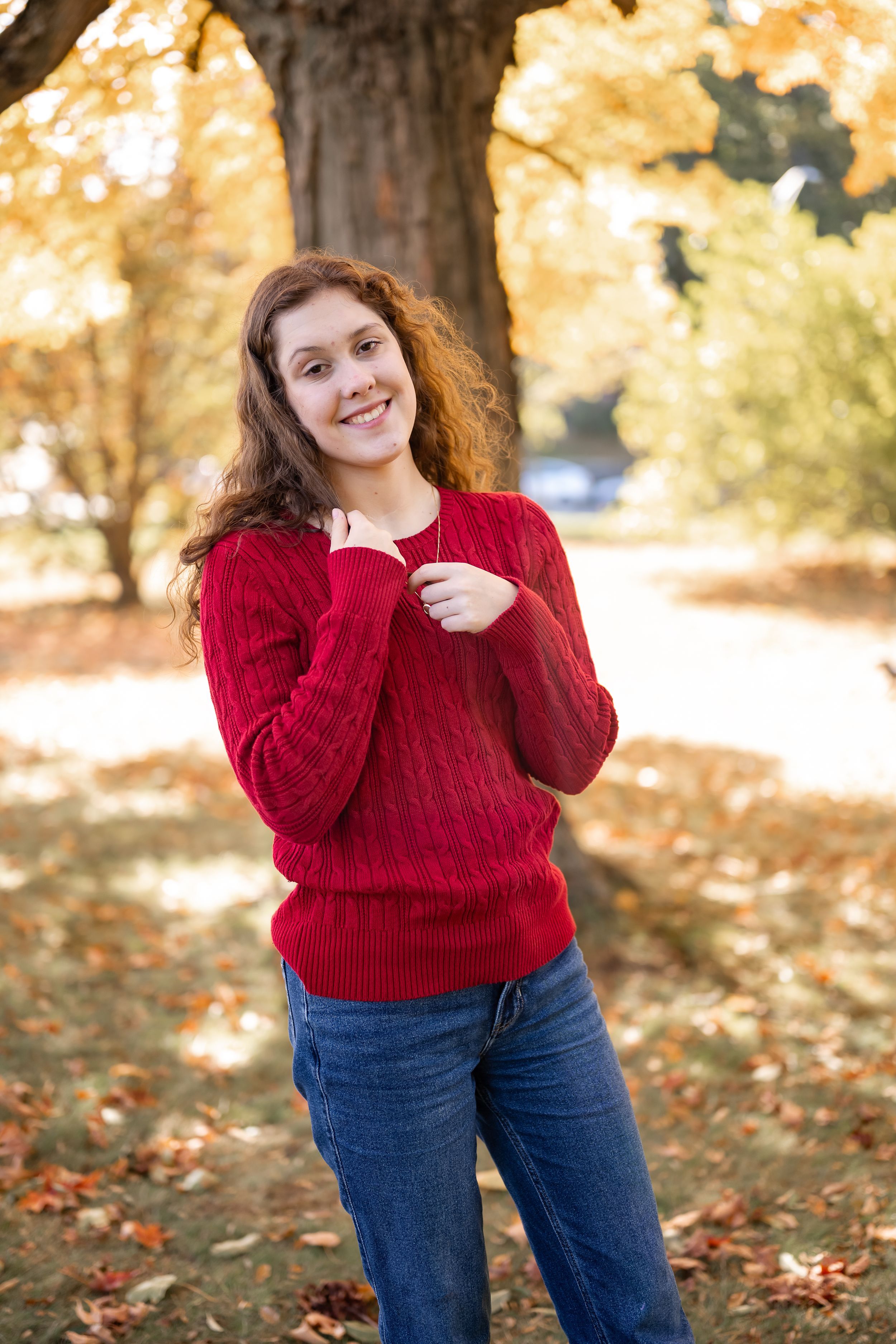 A person in a red cable knit sweater and jeans poses playfully against an autumn backdrop with fallen leaves.