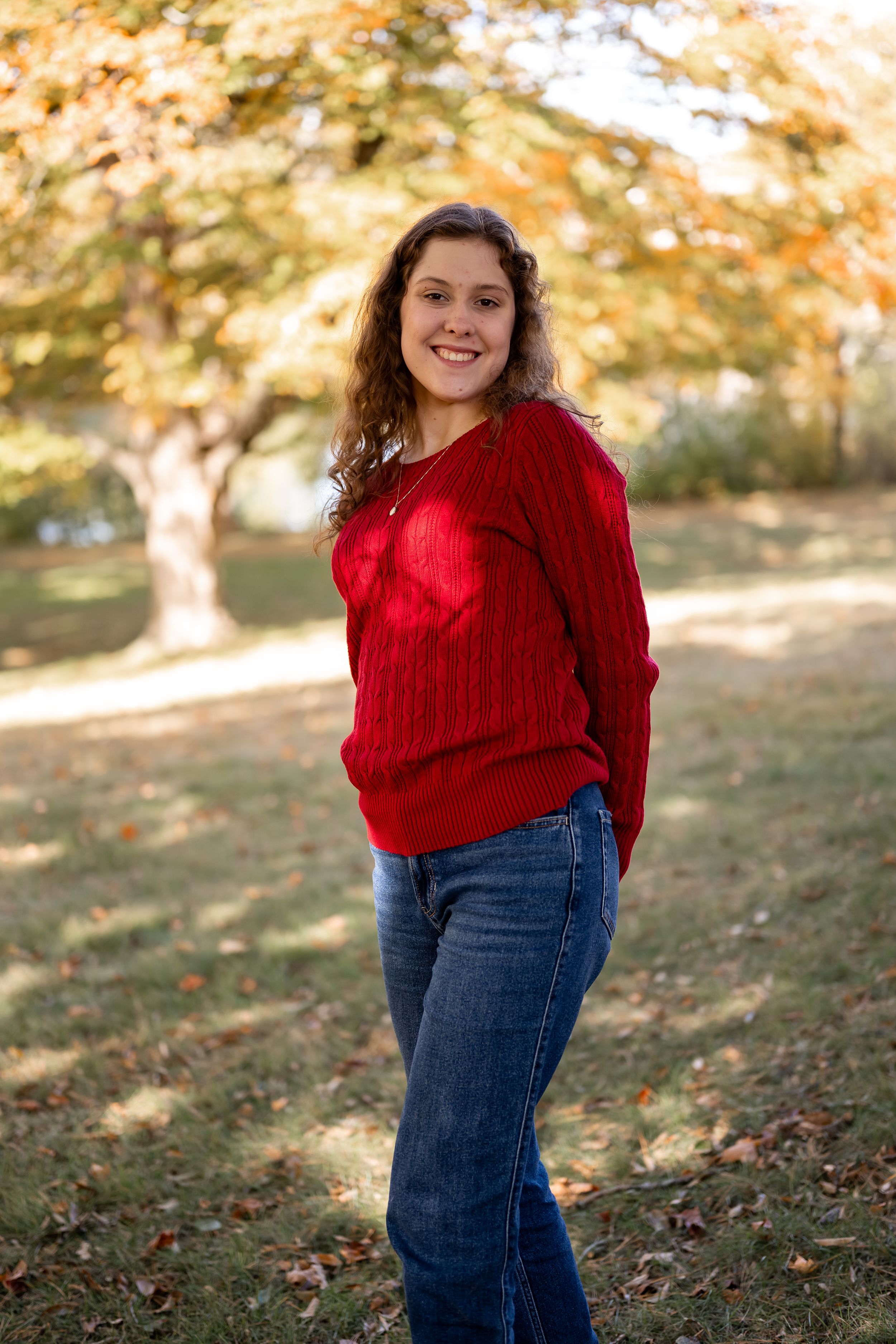 A person wearing a red knit sweater and jeans poses outdoors during autumn with fall foliage in the background.