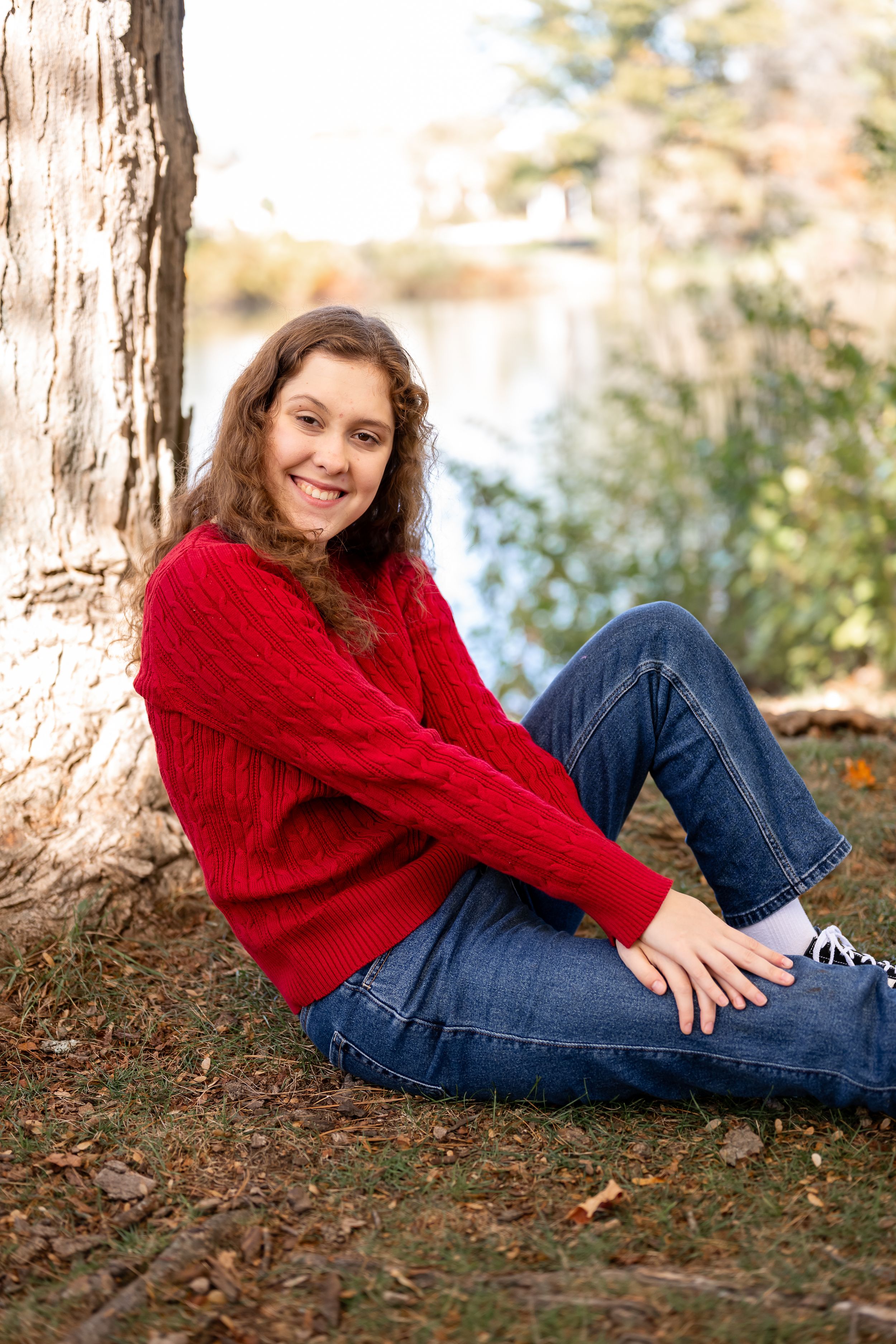 Person in red sweater and blue jeans lounges on autumn ground beneath tall pine trees.