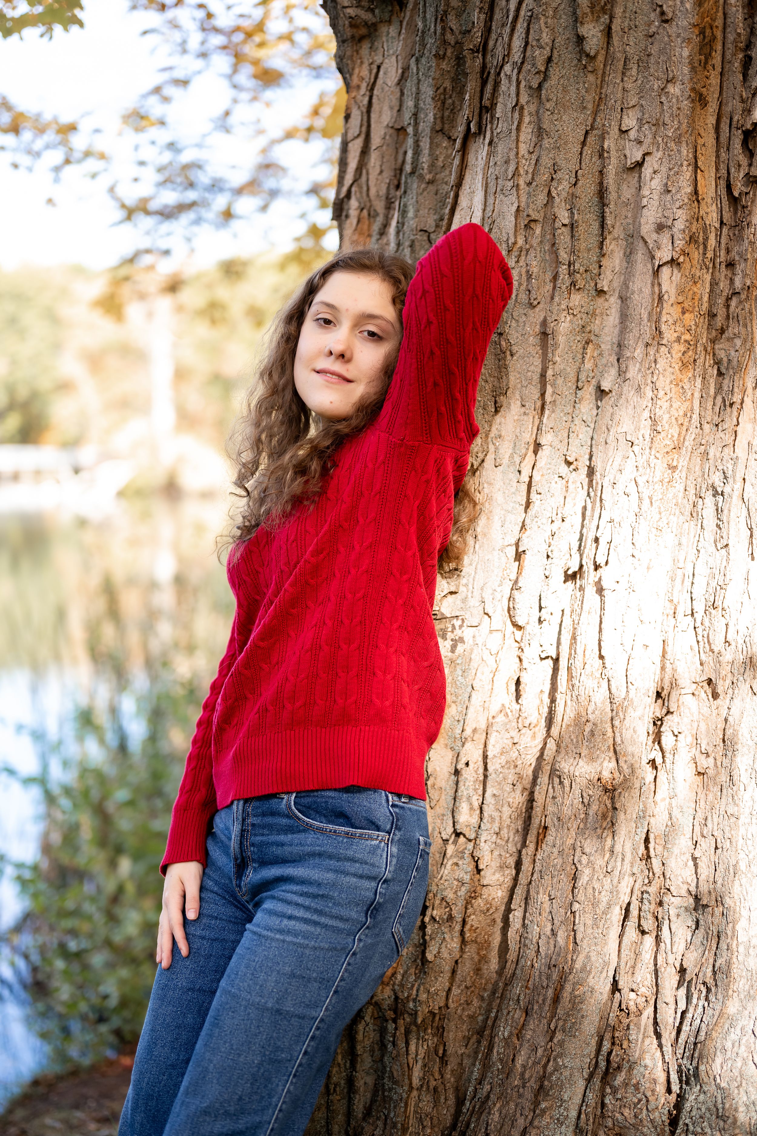 Someone wearing a bright red sweater and blue jeans leans casually against a large tree trunk in a park setting.