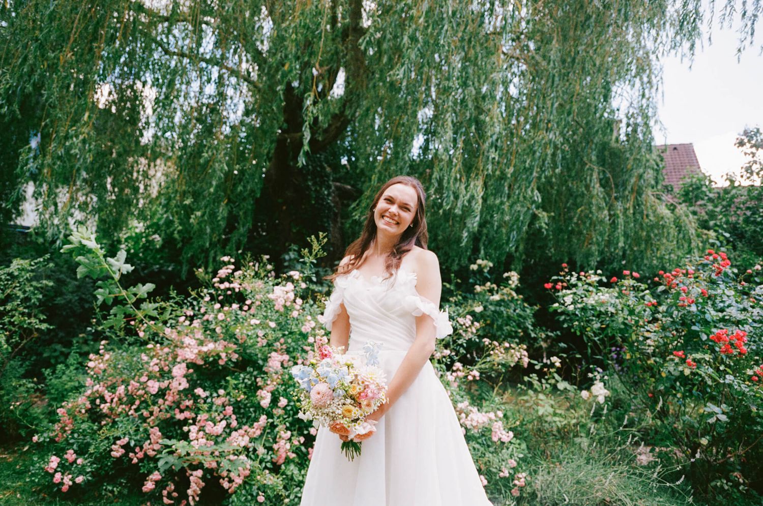 Bride in white gown holding colorful bouquet smiling in lush garden with blooming roses and willow tree.