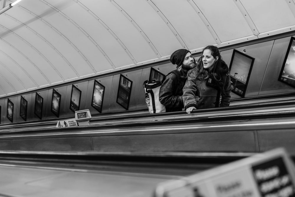 Two people smiling and hugging on escalator on the London Underground station with multiple levels visible in black and white photography.