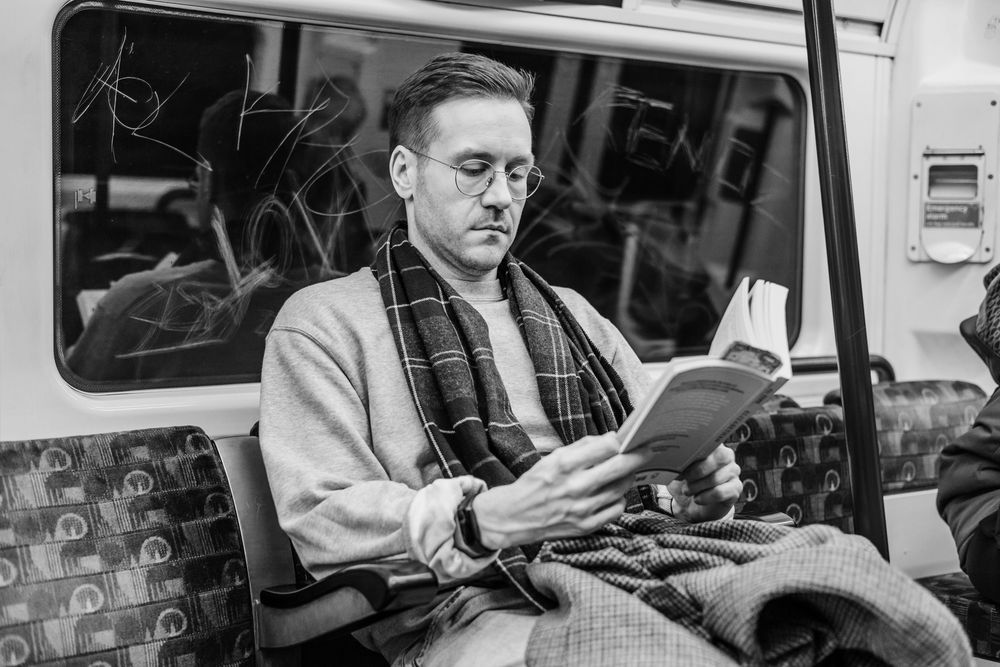 Man with glasses and a scarf reads a novel on the London Underground