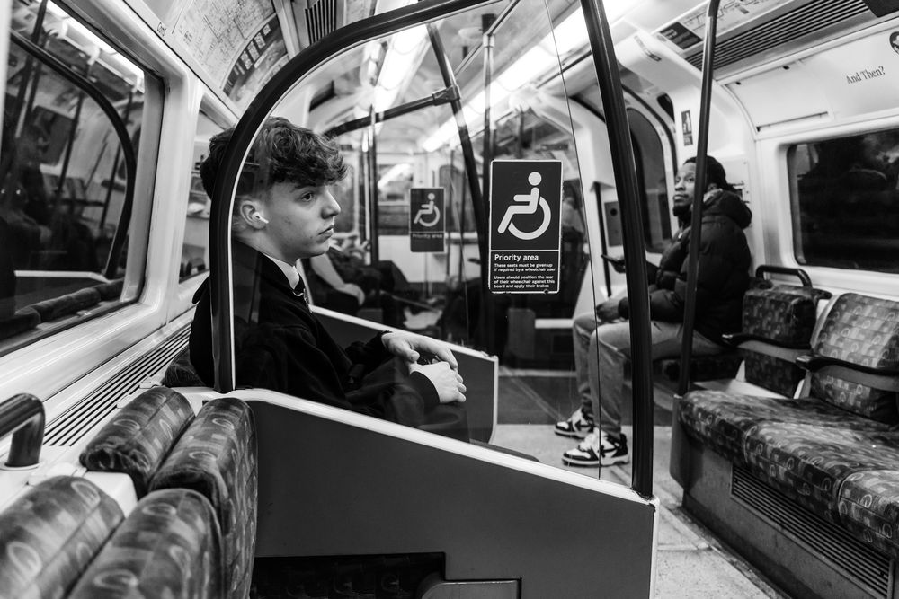 Black and white photo of passengers riding in a London Underground train car with vintage patterned seats and accessibility signage.
