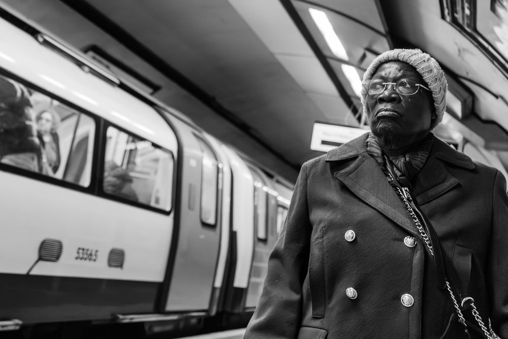 A black and white photograph of a person wearing a coat standing on a subway platform with a train in the background.
