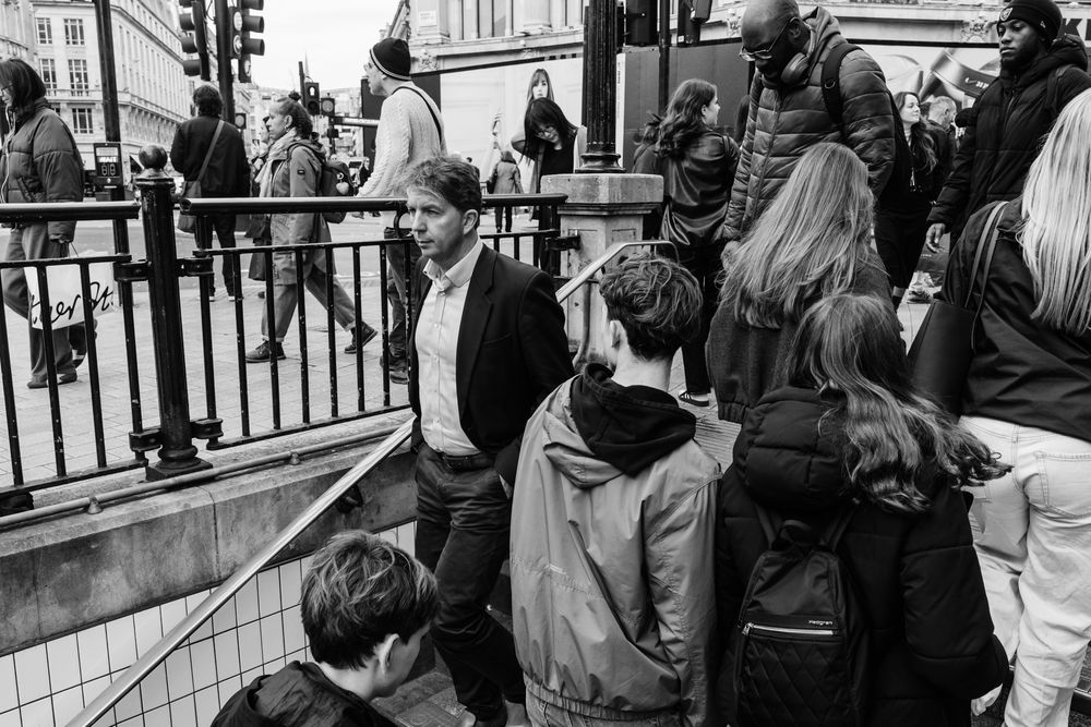 A black and white street scene shows a crowd of people waiting near railings in an urban setting.