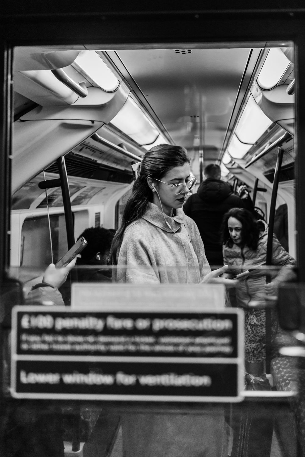 Black and white photograph showing passengers inside a subway car with overhead text signs and lighting fixtures.