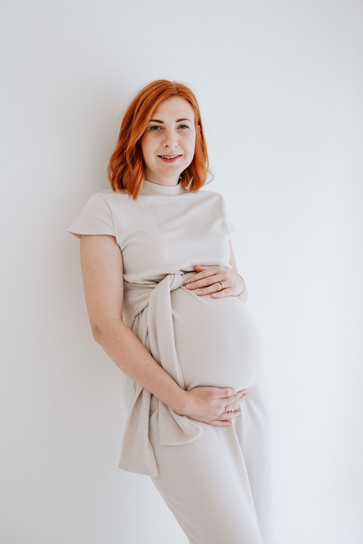 Expecting mother in neutral dress poses against light background at natural light studio maternity photoshoot