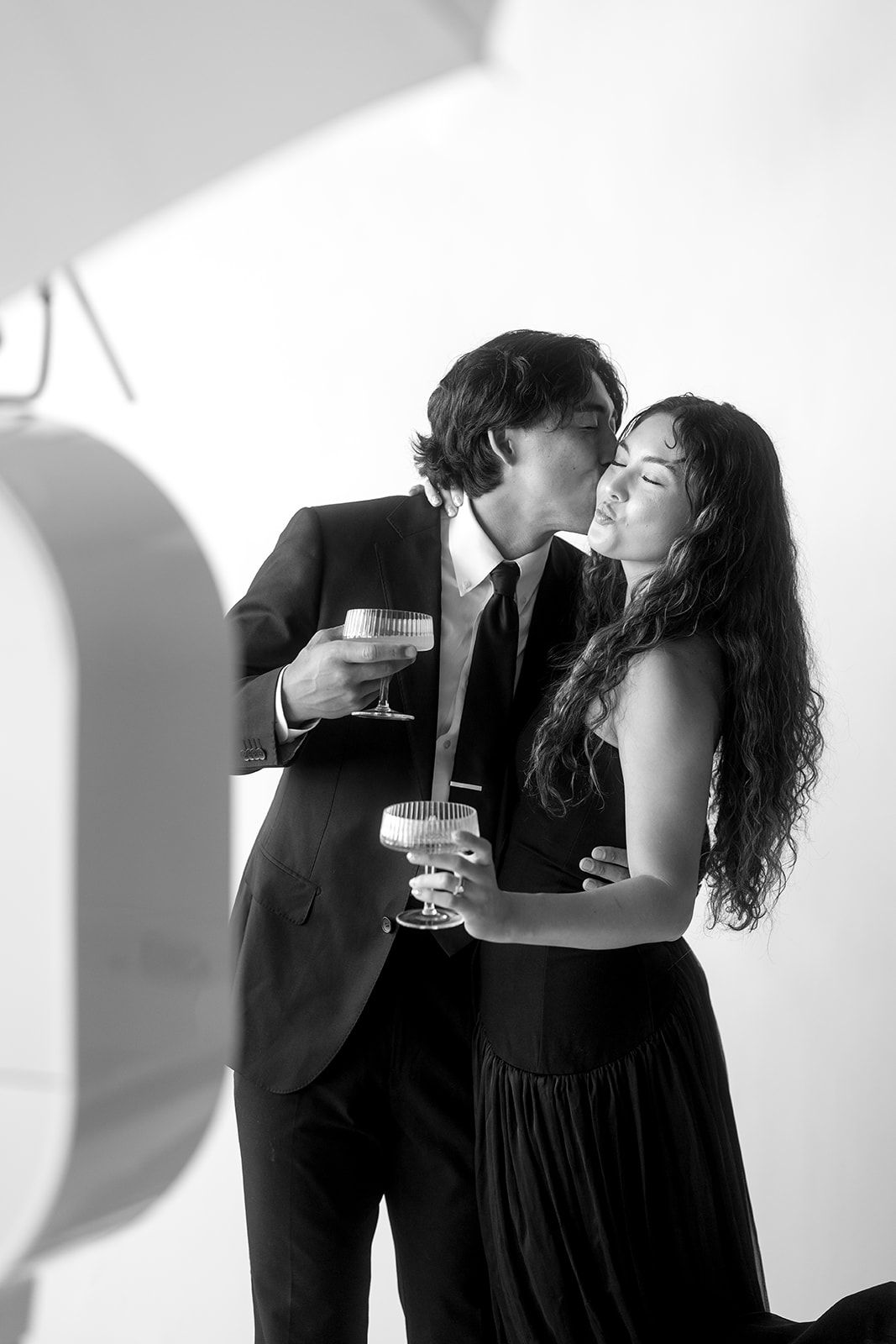 A couple in black formal attire shares a cute moment, captured by a photo booth, while holding wine glasses against a white background.