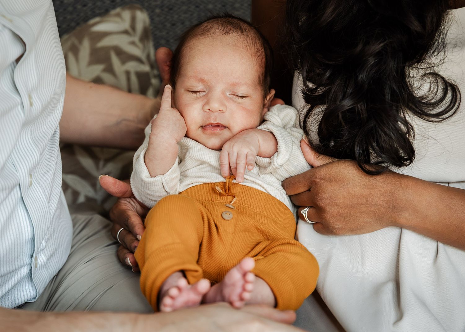 A newborn baby dressed in mustard coloured leggings sleeps peacefully in his parents arms. In home Bristol newborn photography