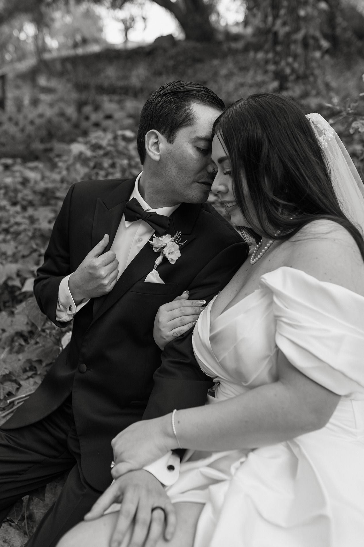 A bride in an off-shoulder white gown and her groom while holding a bouquet of white roses in a wooded setting at The Oaks at Duncan Lane.
