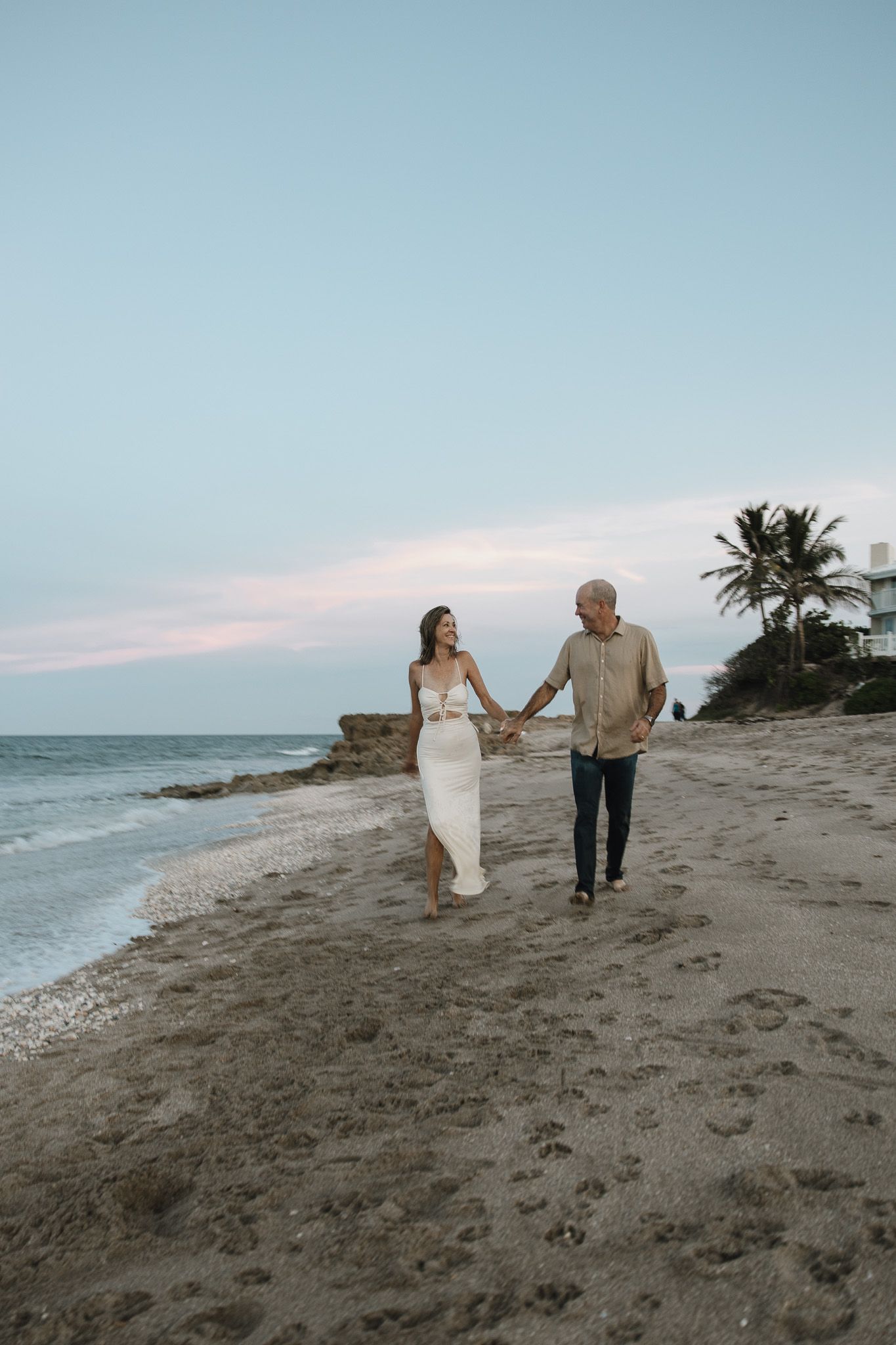A couple walks hand in hand along a serene beach at sunset with palm trees and ocean waves in the background.